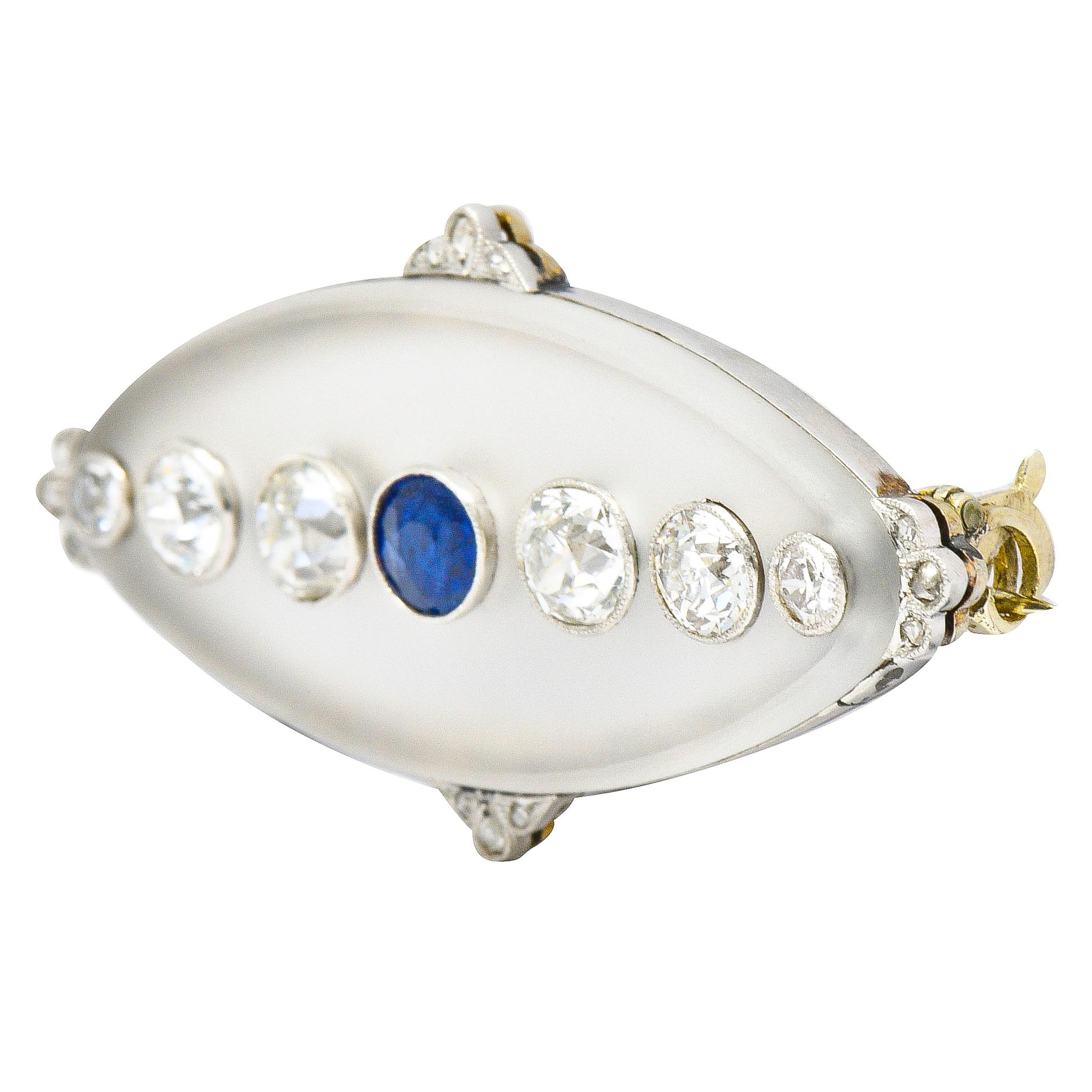 Elongated oval brooch is comprised of frosted camphor glass measuring approximately 37.0 x 15.5 mm

Centering a bezel set round cut sapphire, bright blue in color, and weighs approximately 0.38 carat

Flanked by old European cut diamonds weighing in