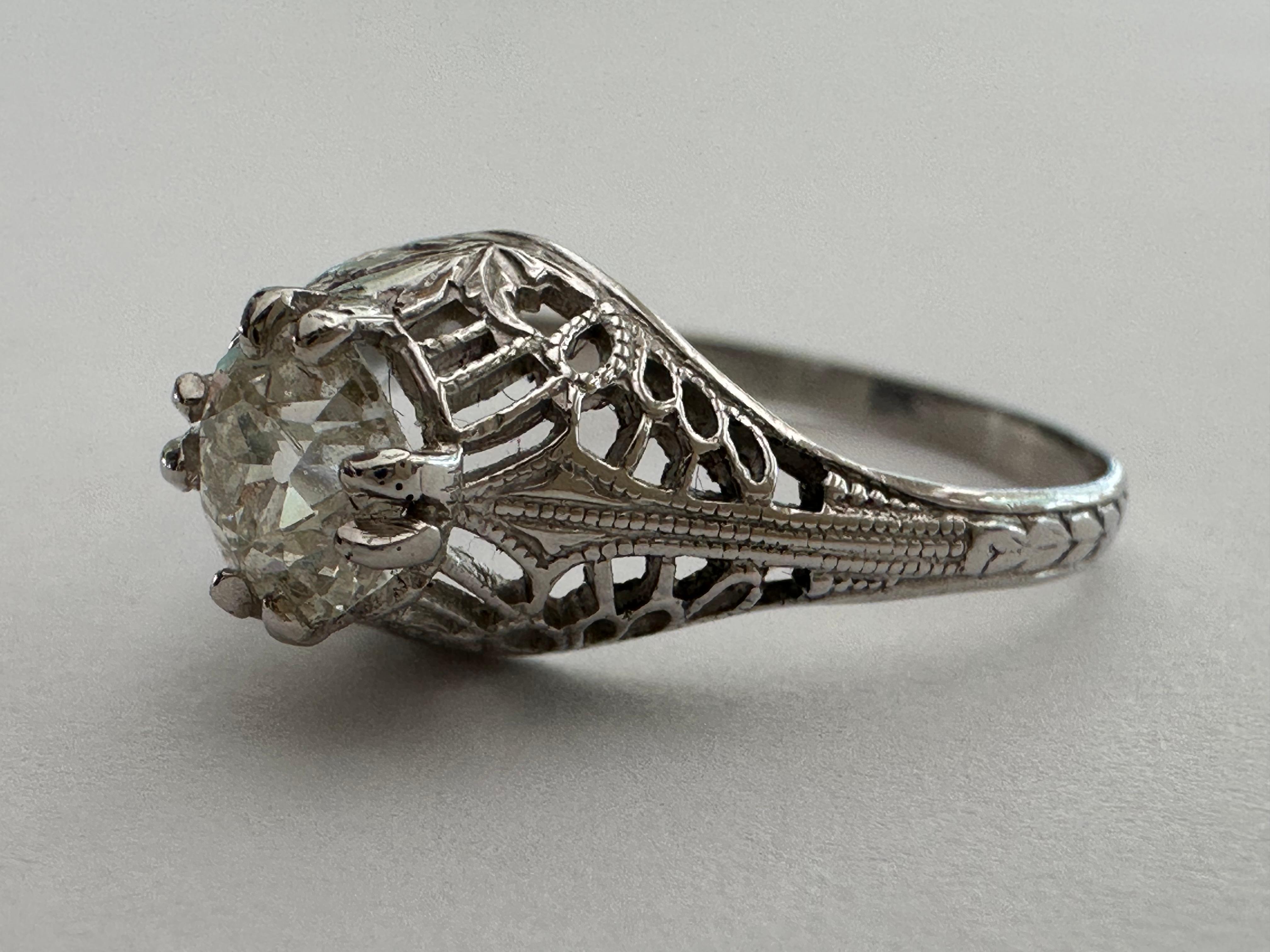 Una piedra central de diamante de talla europea antigua de aproximadamente 0,63 quilates, color I y claridad SI, brilla con luz propia en este anillo solitario de estilo Art Decó tardío sobre una graciosa montura de filigrana adornada con detalles