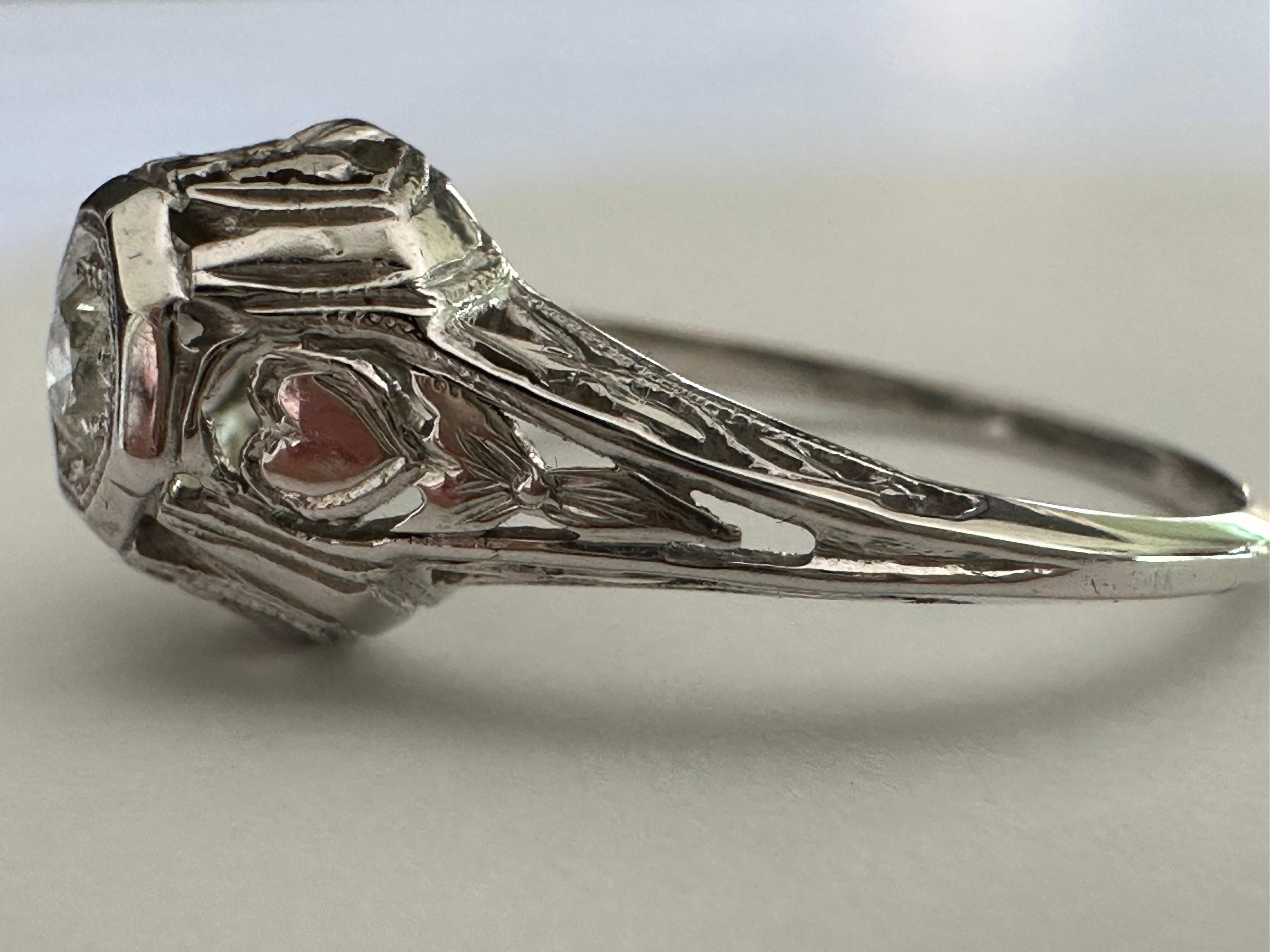 Una piedra central de diamante de talla europea antigua de aproximadamente 0,20 quilates, color F-G y claridad VS, brilla con luz propia en este anillo solitario Art Decó, sobre una graciosa montura de filigrana adornada con corazones y pequeñas