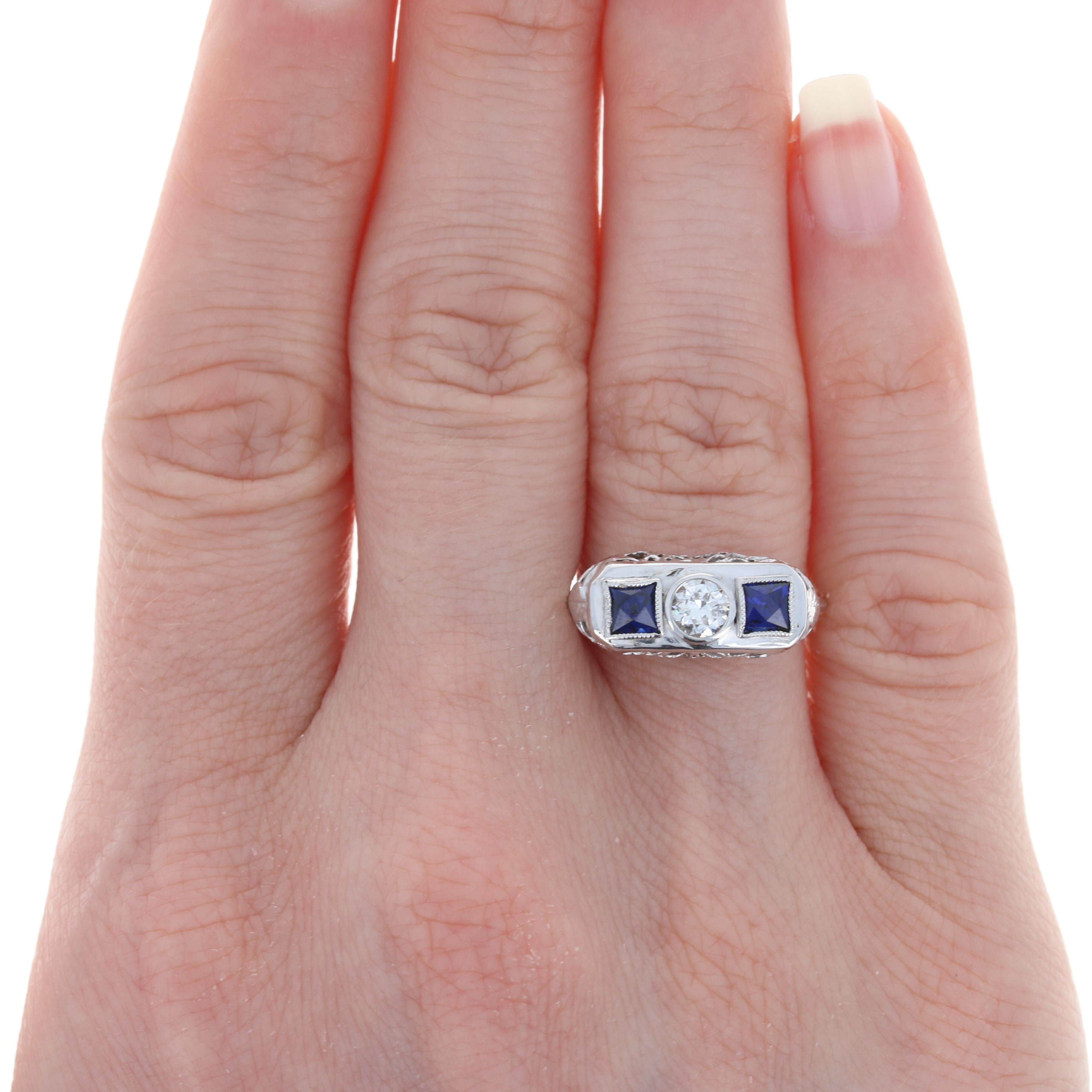 Sheer Art Deco allure! Crafted in the 1920s - 1930s, this luxurious 18k white gold vintage ring showcases a sparkling diamond solitaire set between two synthetic blue sapphires which are highlighted by ornate filigree and milgrain accents to create