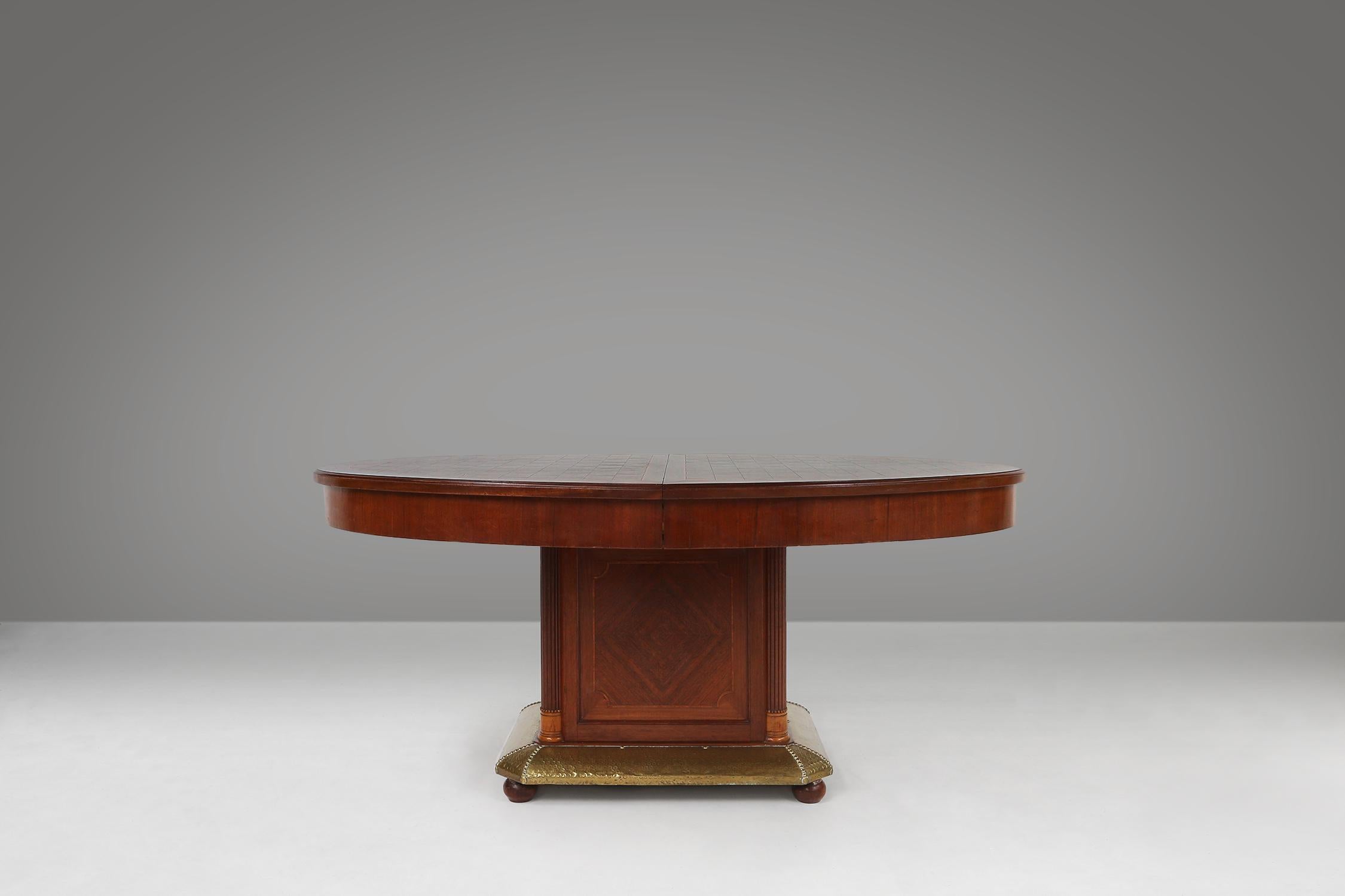 This Art Deco dining table was made by De Coene in Belgium in 1930. This table is made of wood with inlaid wood and has a brass base. The table is oval in shape with a chessboard pattern on the top. The base is square with a geometric design. The