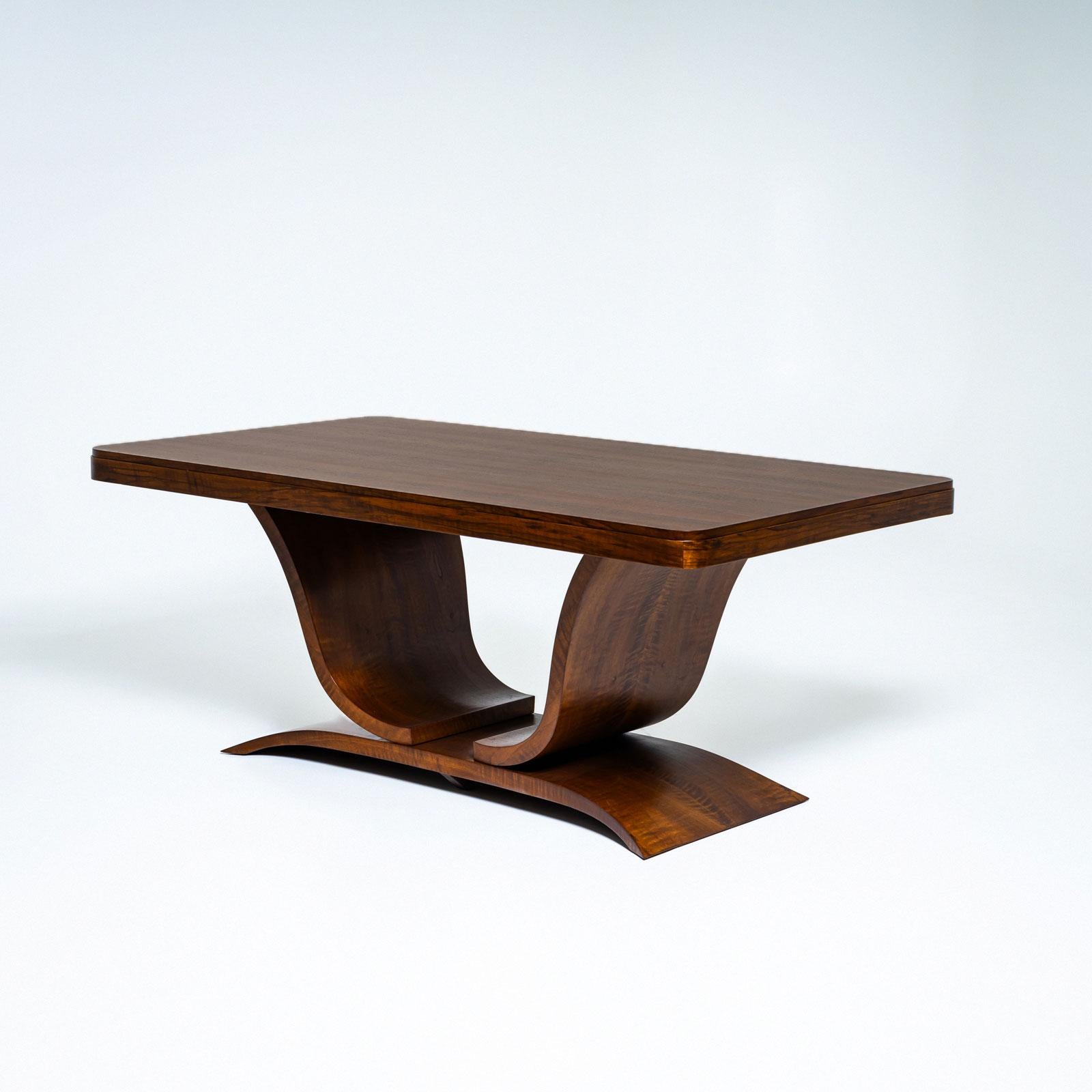 An elegant dining table featuring a tulip-shaped base resting on a curved rectangular plinth. The rectangular tabletop, with rounded corners and a simply stepped edge, showcases a stunning walnut veneer pattern. The table has been professionally