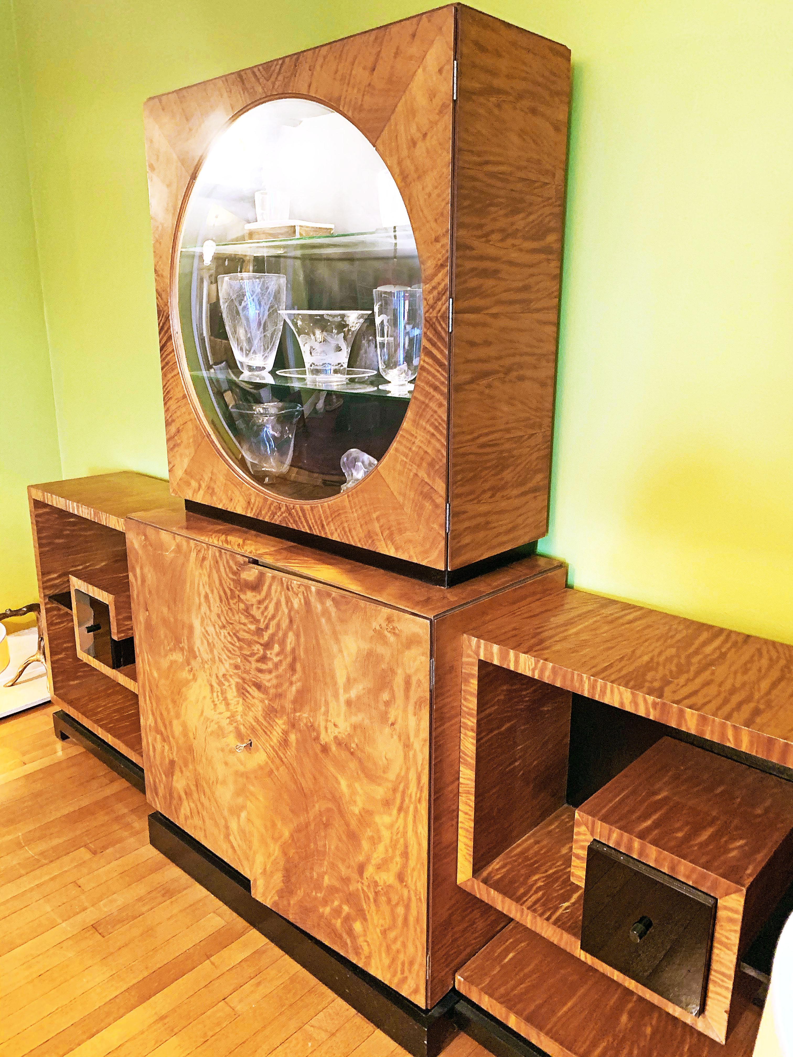 Stunning in concept and execution, this large, impressive Moderne-Art Deco cabinet consists of four parts, all centered around an internally-lit porthole vitrine with glass shelves and convex window, all finished in a golden flame mahogany that has