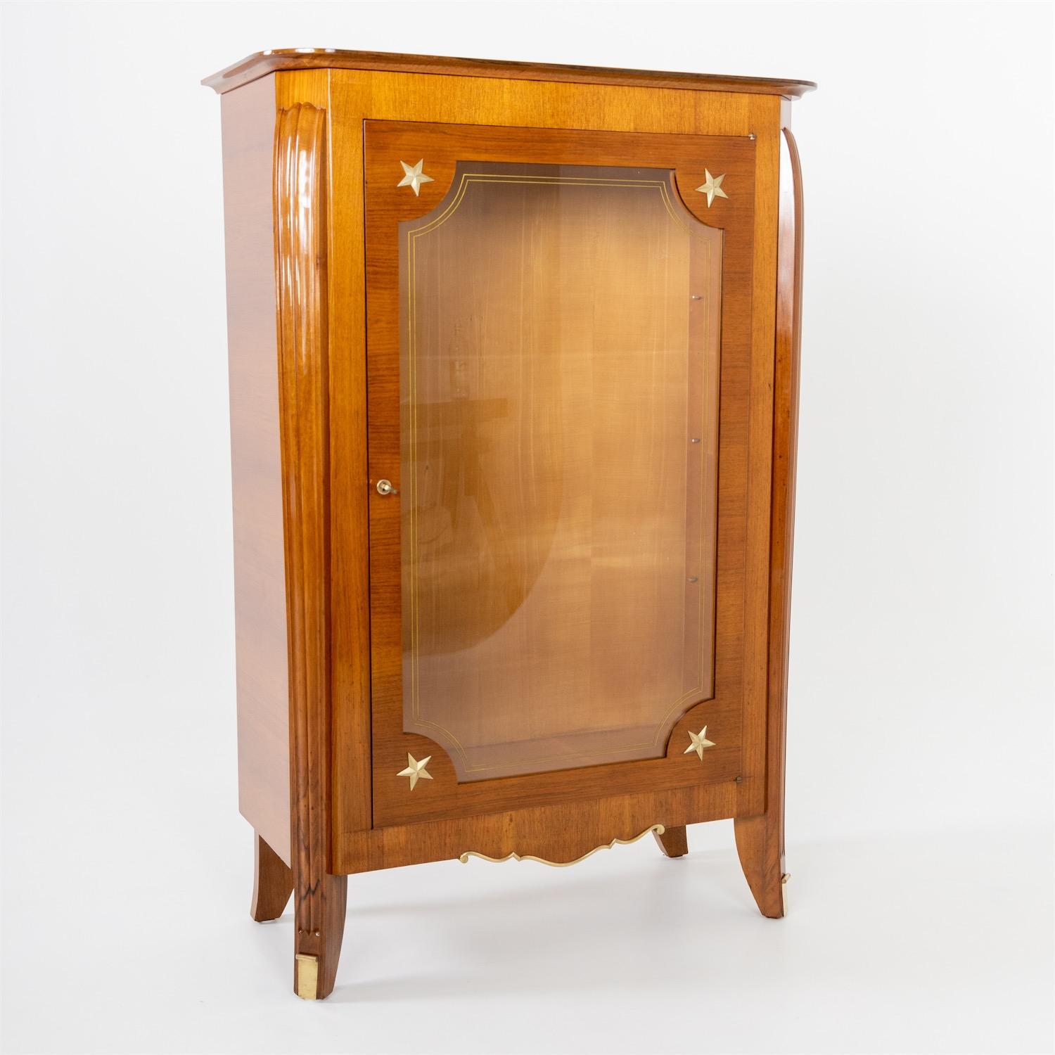 Single door glazed display case with brass star-shaped appliques and slightly trapezoidal body. The pilasters are fluted and slightly shouldered.