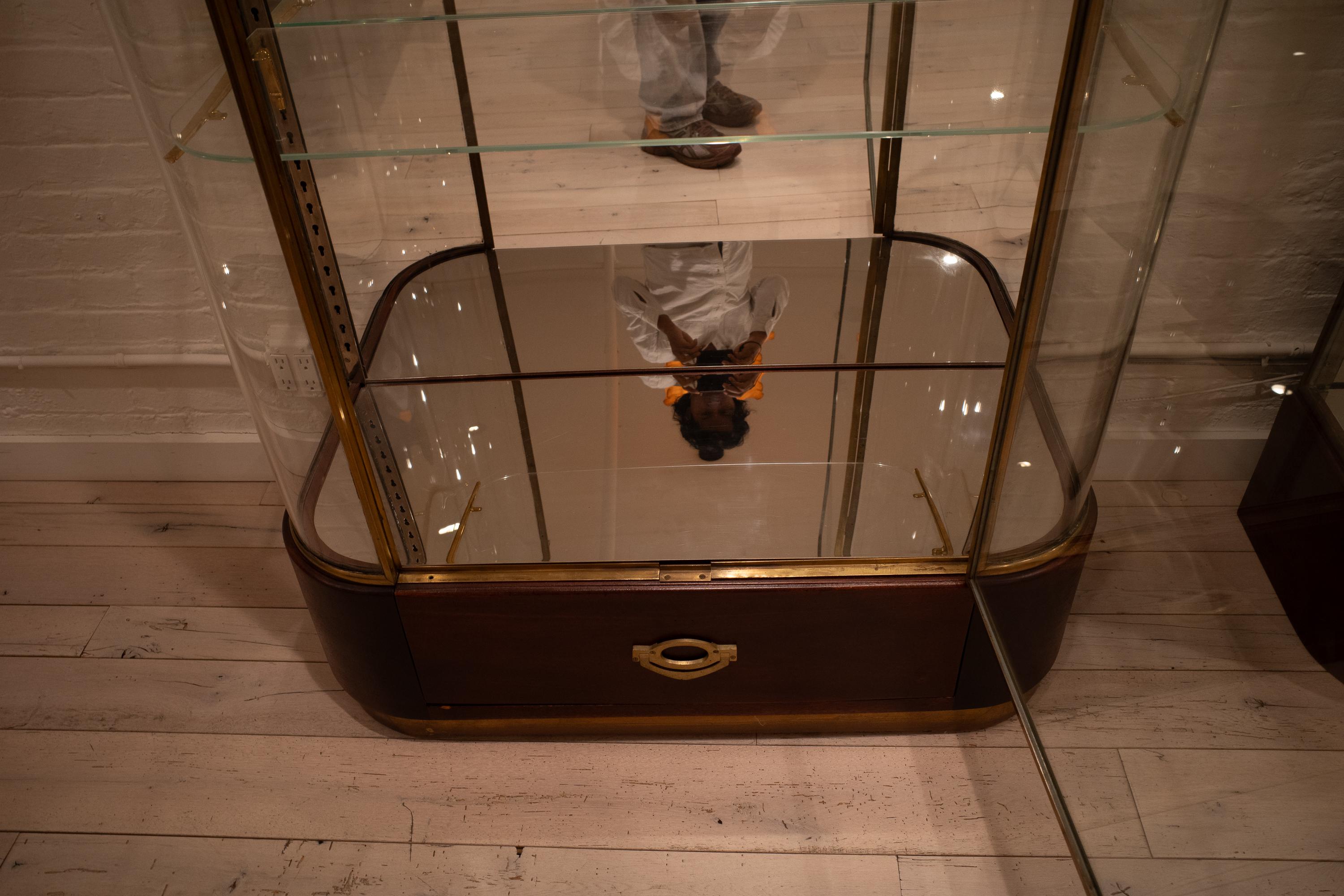 This art deco era display cabinet, circa 1930, was made in Paris by LENA at 160 Rue Oberkampf. Constructed from brass and mahogany, it features a large glass door that opens to reveal adjustable glass shelves with brass supports and a mirrored