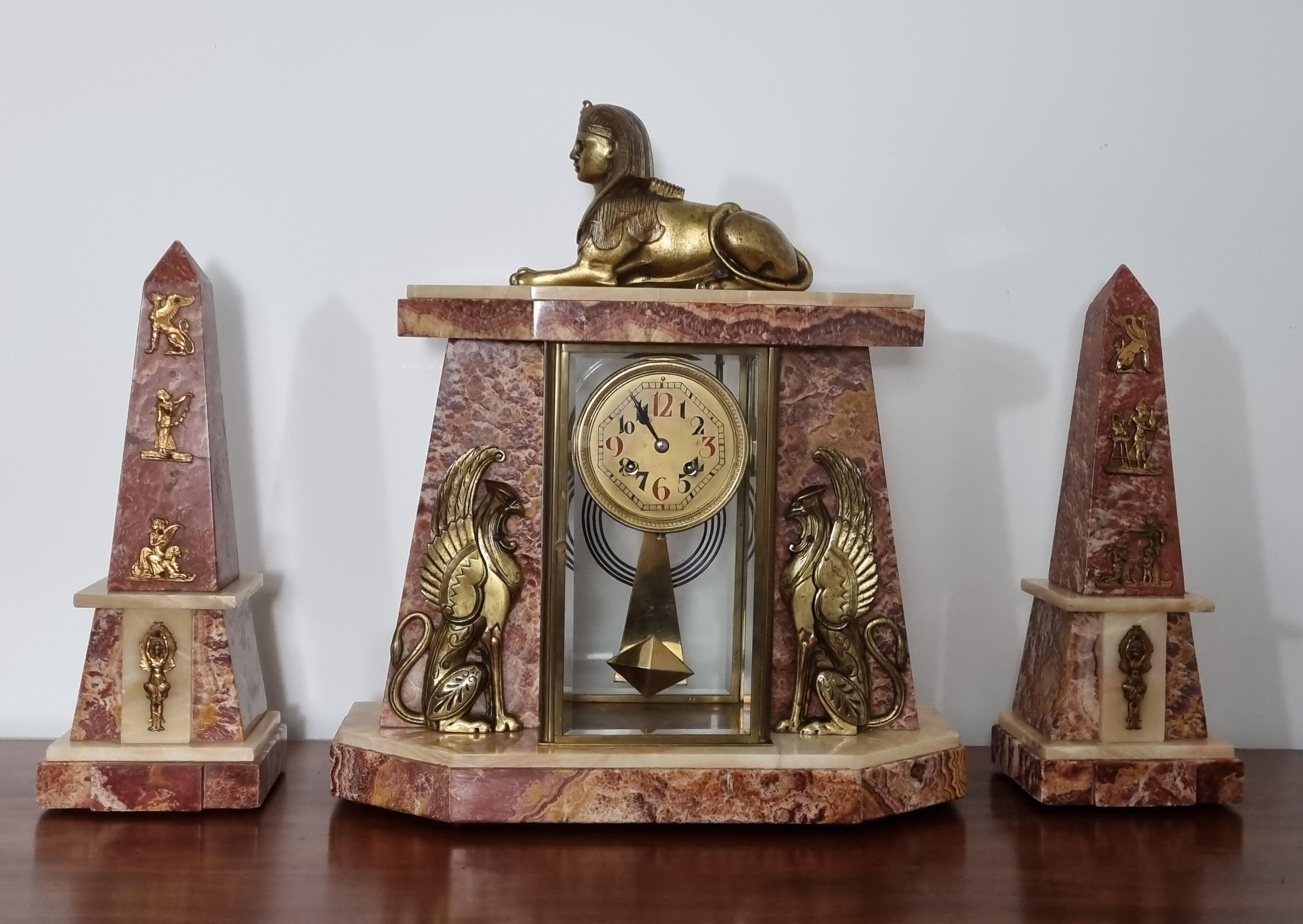 A stylish Art Deco Egyptian Revival clock set featuring a central timepiece flanked by a pair of obelisks, this piece exudes the grandeur and mystique of the early 20th-century fascination with ancient Egypt. Atop the clock sits a regal sphinx cast
