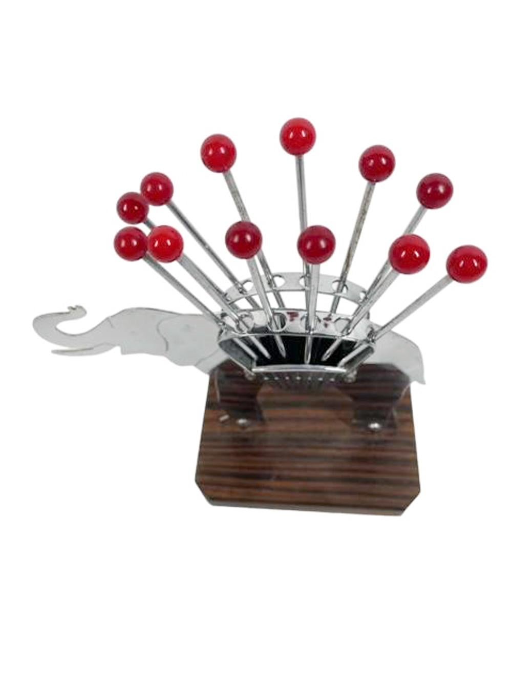 Art Deco cocktail pick set in chrome and wood with red ball topped picks with forked tips. Two-dimensional chromed elephant figure standing on a   striped wood base carrying a pair of baskets, each holding six picks.