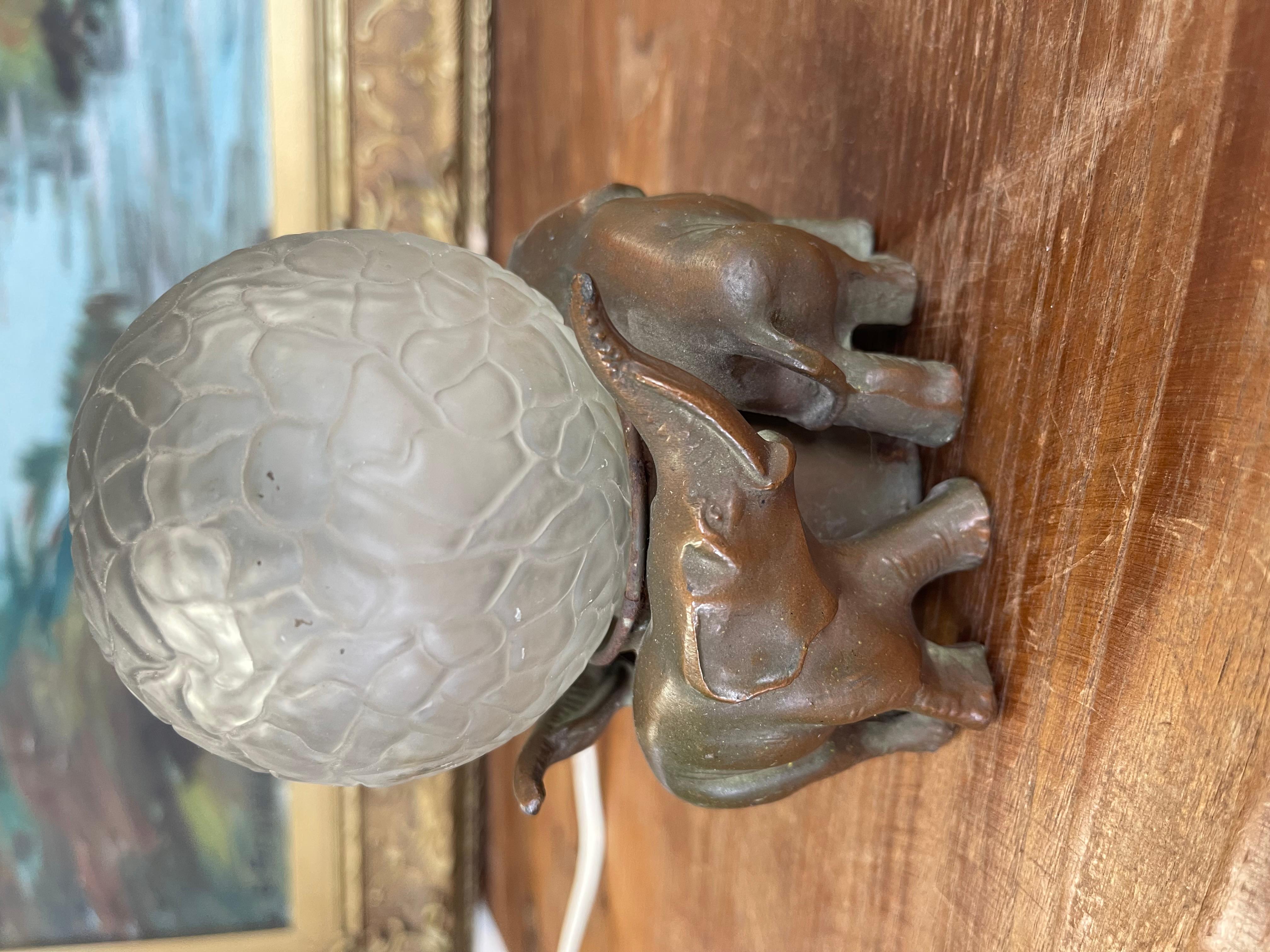 A charming Art Deco era accent lamp featuring a trio of elephants supporting a frosted globe shade. The elephants are finished in a warm bronze-toned patina, their trunks raised around the central socket. The textured frosted globe is patterned with