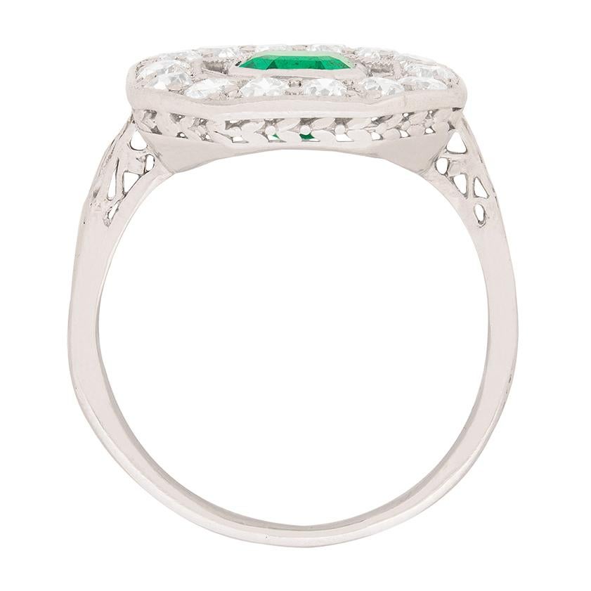This beautiful dress ring dates back to the 1920s. It features a deep green emerald within a rub over setting weighing 0.92 carat. It is a natural emerald and has been beautifully haloed by old cut diamonds. Each of the diamonds weighs 0.05 carat