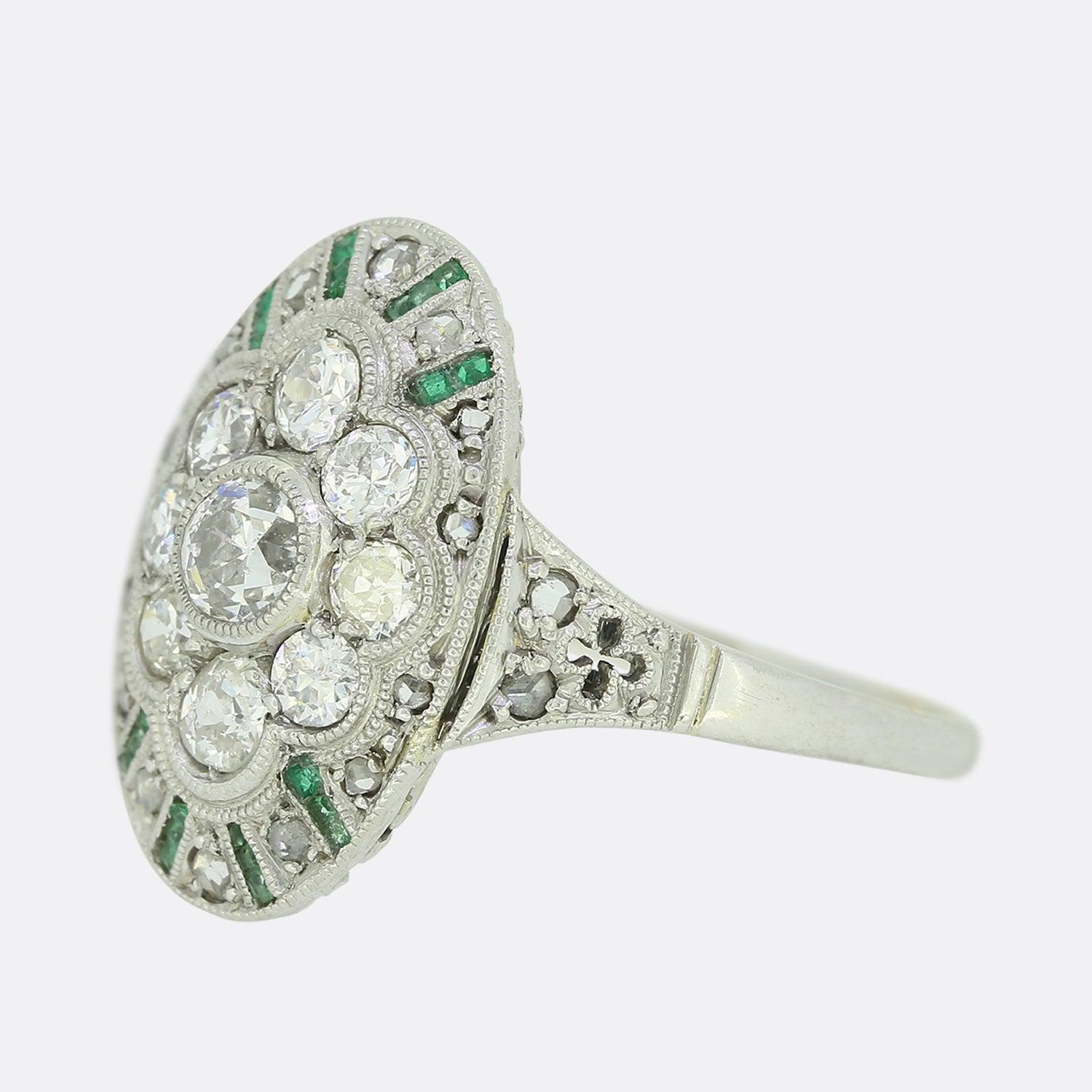 This is a wonderful emerald and diamond cluster ring from the Art Deco era. The ring features nine central old cut diamonds in a cluster formation and surrounded by calibre cut emeralds and rose cut diamonds in an oval shaped 18ct white gold