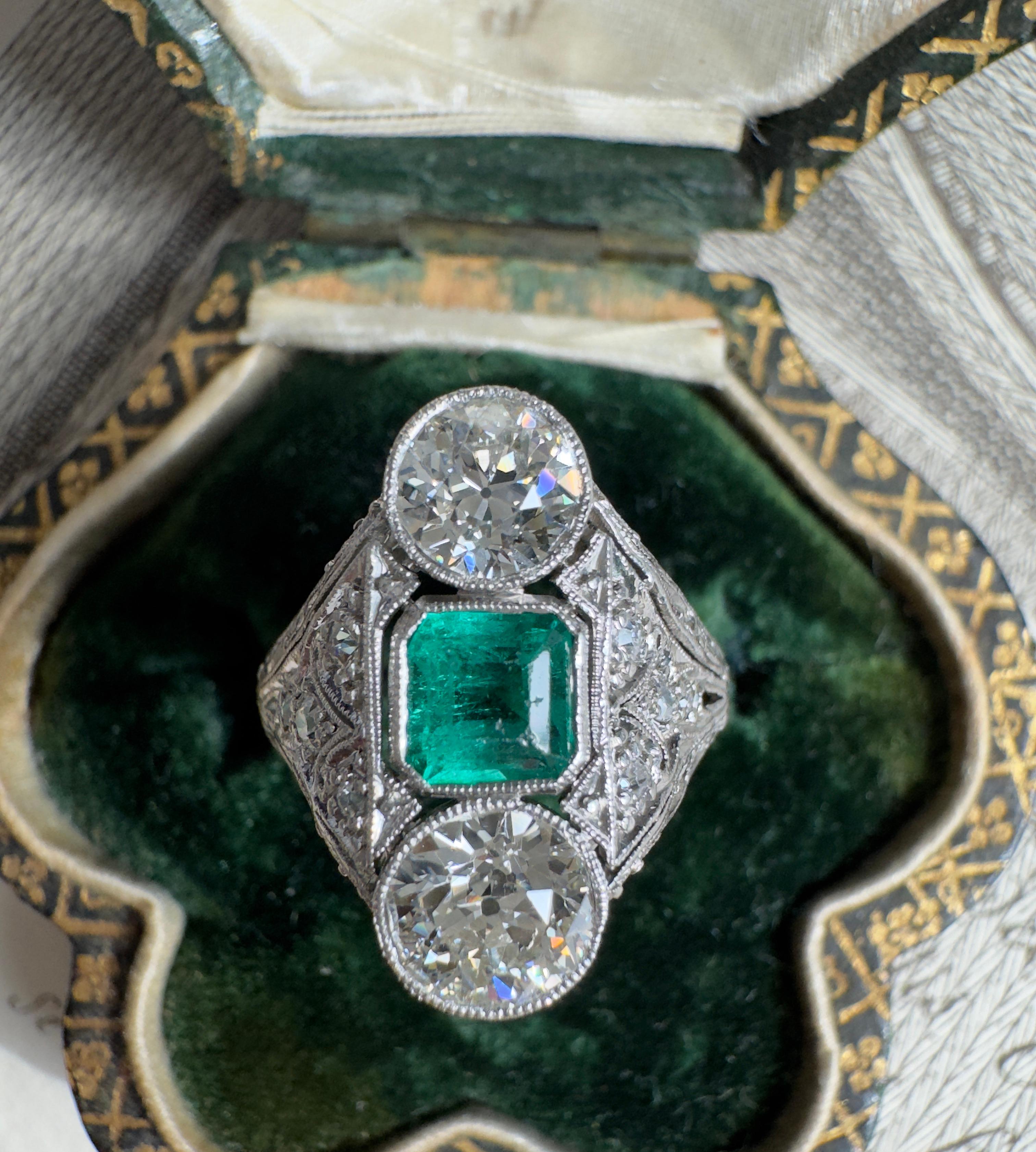 This charming early Art Deco ring centers on a glowing Colombian emerald, weighing .60 carat, embraced by a pair of sparkling European-cut diamonds. Low profile and finger hugging, the platinum setting frames the trio with soft shoulders aglitter