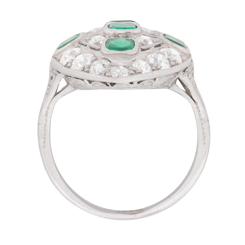 Five old cushion cut emeralds diamonds in a bright shade of green comprise the horizontal and vertical centre of this original Art Deco dinner ring.

Millegrained openwork detailing separates the emeralds, which are rub over set in box collets and