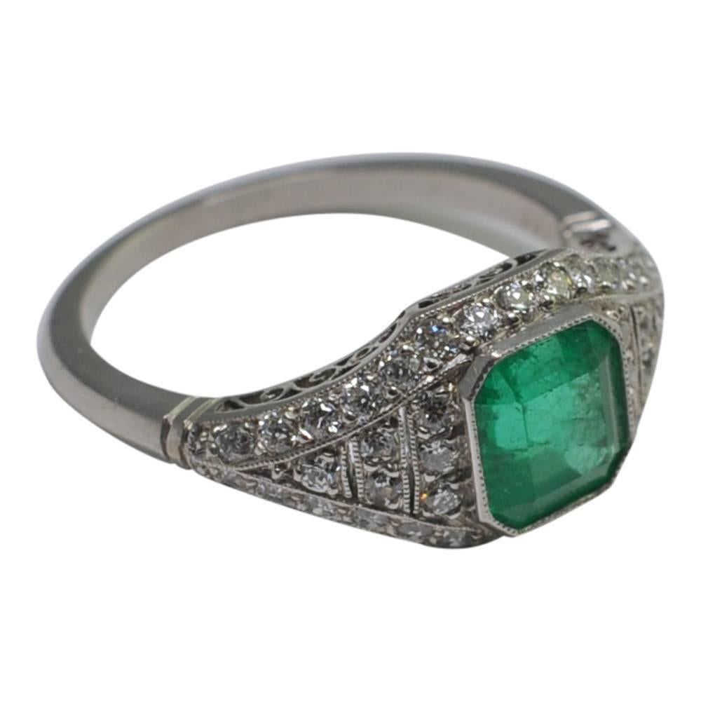 Art Deco emerald and platinum vintage engagement ring; this is formed of a curved hoop over a pierced gallery and is bezel set with a vibrant green step cut emerald weighing 1.26ct, which is surrounded by Old European Cut diamonds totalling 0.84ct
