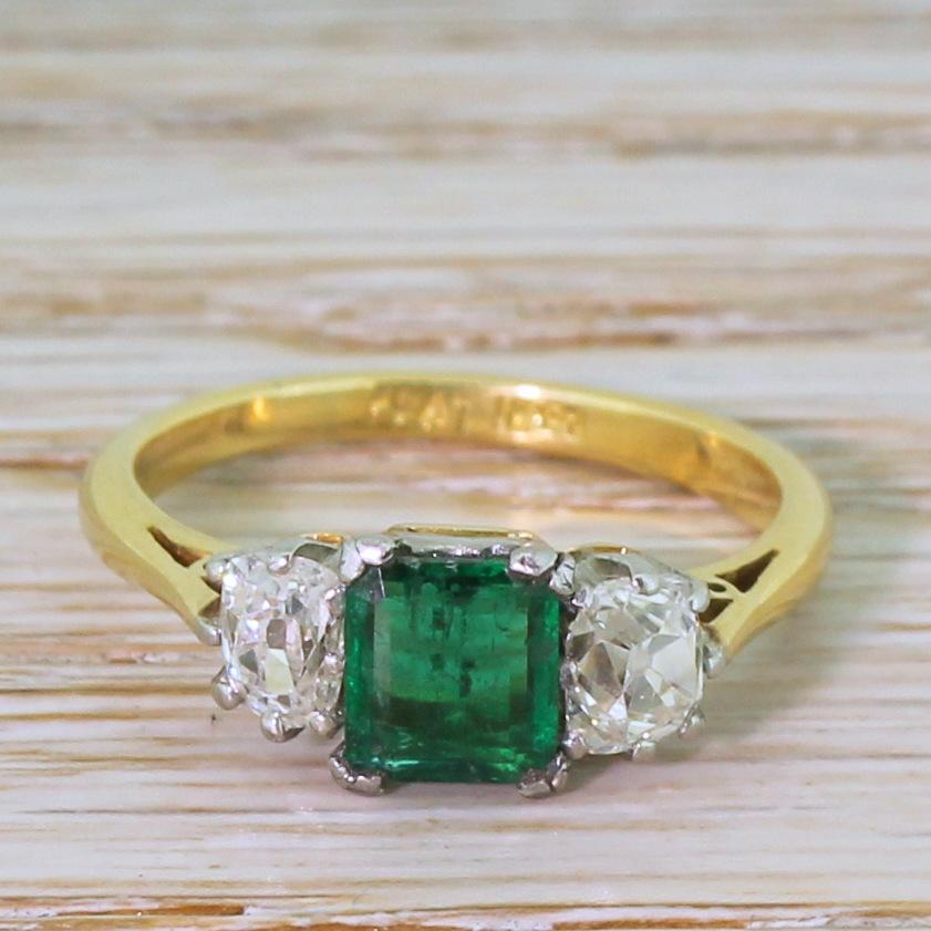 A perfectly balanced emerald and diamond trilogy ring. The square step cut emerald in the centre displays a bright, leafy green and is secured in a platinum four-claw “H” collet. A pair of white, bright and clean old mine cut diamonds are set in
