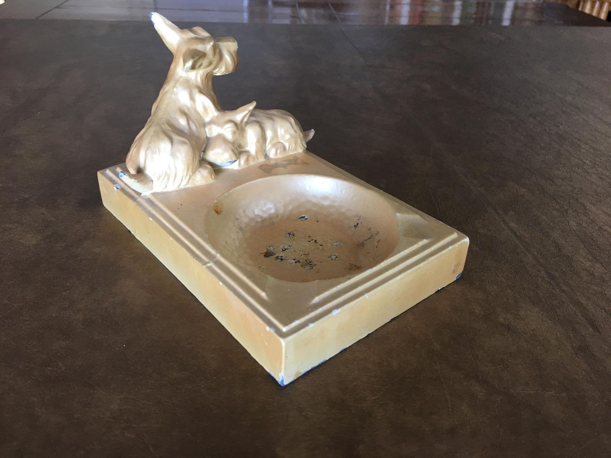 Cendrier Art Déco en métal émaillé beige en forme de chien terrier avec porte-cigarette et 2 chiens sculptés sur le dessus. Cette pièce présente une certaine usure dans le bol du cendrier et le long de ses bords.

Dimensions :
4