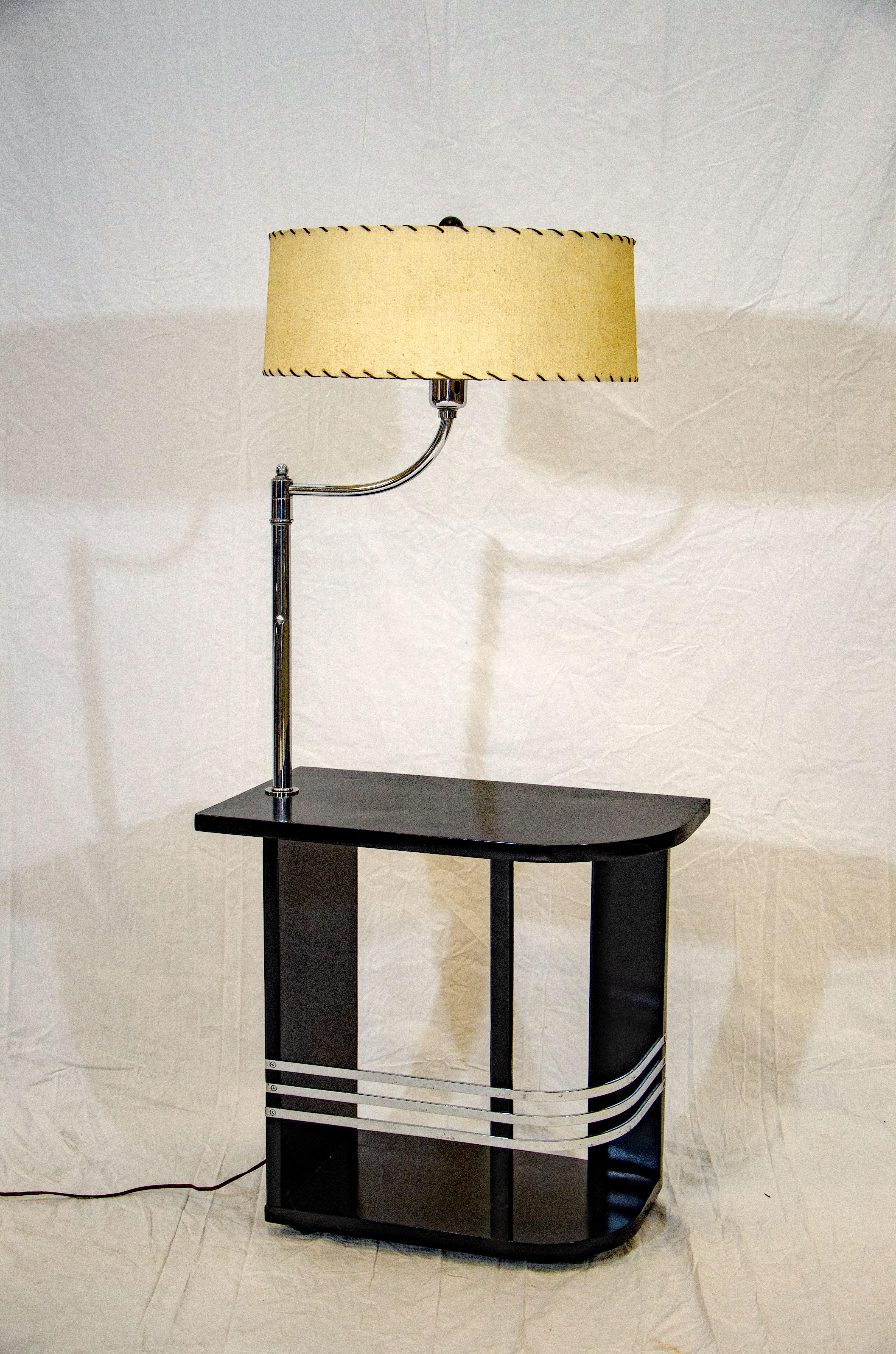 This nice Art Deco end table with a black finish is accented with three curved chrome bands and a chrome lamp. The tabletop is curved at one end. The lamp has a conical milk glass shade which holds a vintage circular shade with black lacing and a