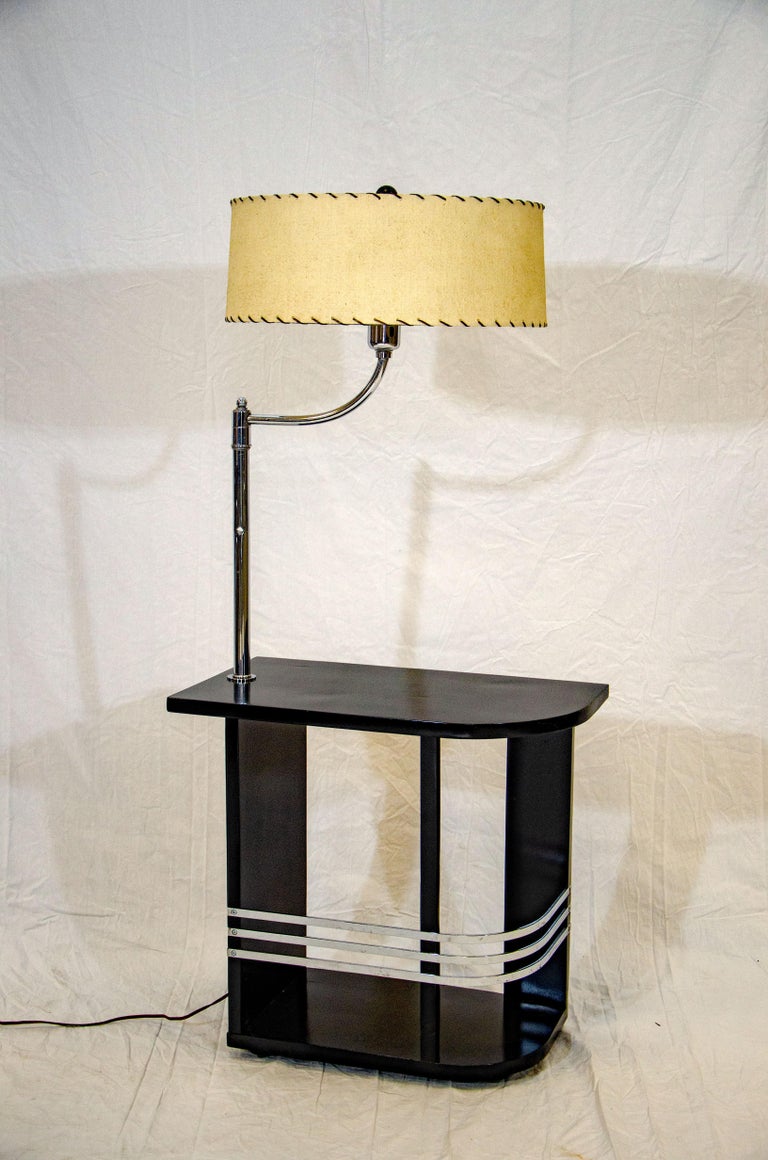 This nice Art Deco end table with a black finish is accented with three curved chrome bands and a chrome lamp. The tabletop is curved at one end. The lamp has a conical milk glass shade which holds a vintage circular shade with black lacing and a