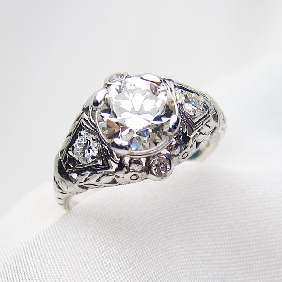This opulent Art Deco platinum filigree ring features a gorgeous transitional-cut diamond weighing 1.74 carats, with a VS2 clarity and J color. The exquisite setting features engraved details accented with four bezel- and bead-set round