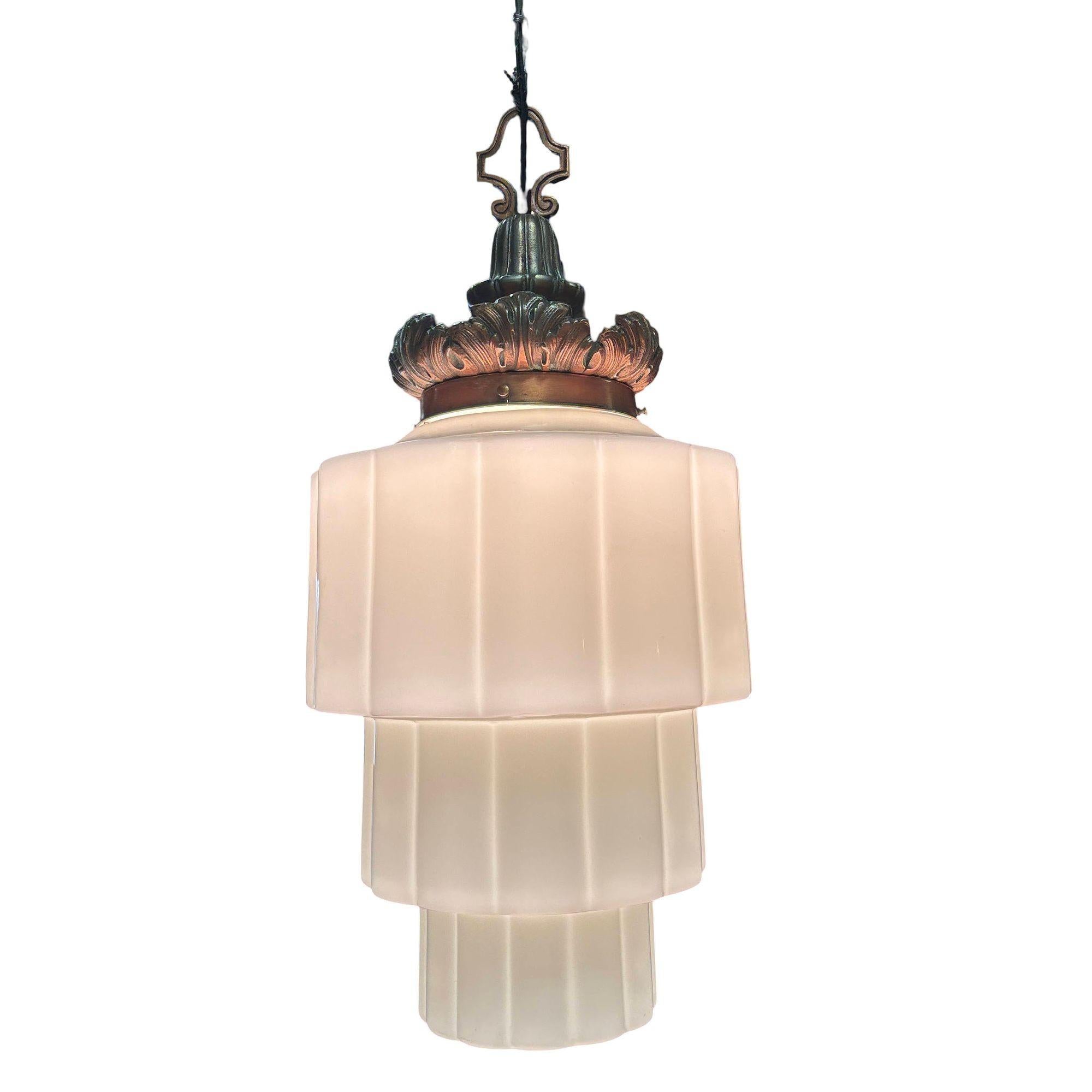 Questa lampada a sospensione Art Deco degli anni '30 presenta un paralume in vetro lattiginoso Art Deco a strati geometrici ed è abbinata a un montatore in ottone fuso con finitura in bronzo caldo. La corona presenta dettagli a foglia d'acanto che
