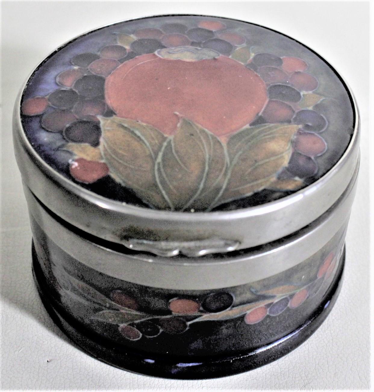 Cette jarre ou boîte de rangement en poterie d'art a été fabriquée par la société Moorcroft Pottery d'Angleterre vers 1935 et réalisée dans leur motif de glaçure grenadine. Cette jarre circulaire a un couvercle à charnière avec une monture en métal