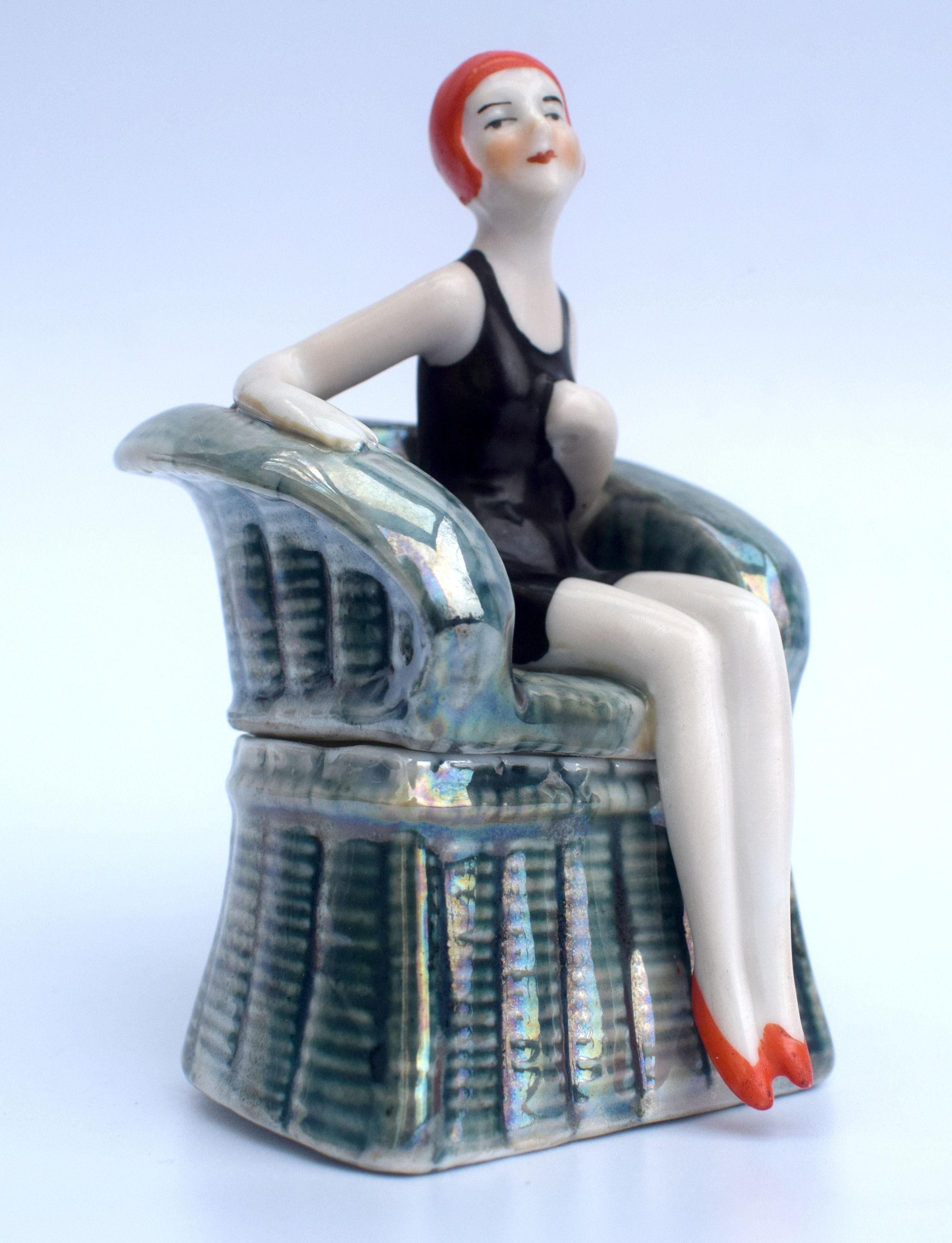 Striking and quite rare is this 1930s Art Deco porcelain powder or trinket box which depicts a stylized female swimmer in the costume of the day sat in a high back wicker chair, she would of been holding an umbrella. Beautiful coloring and