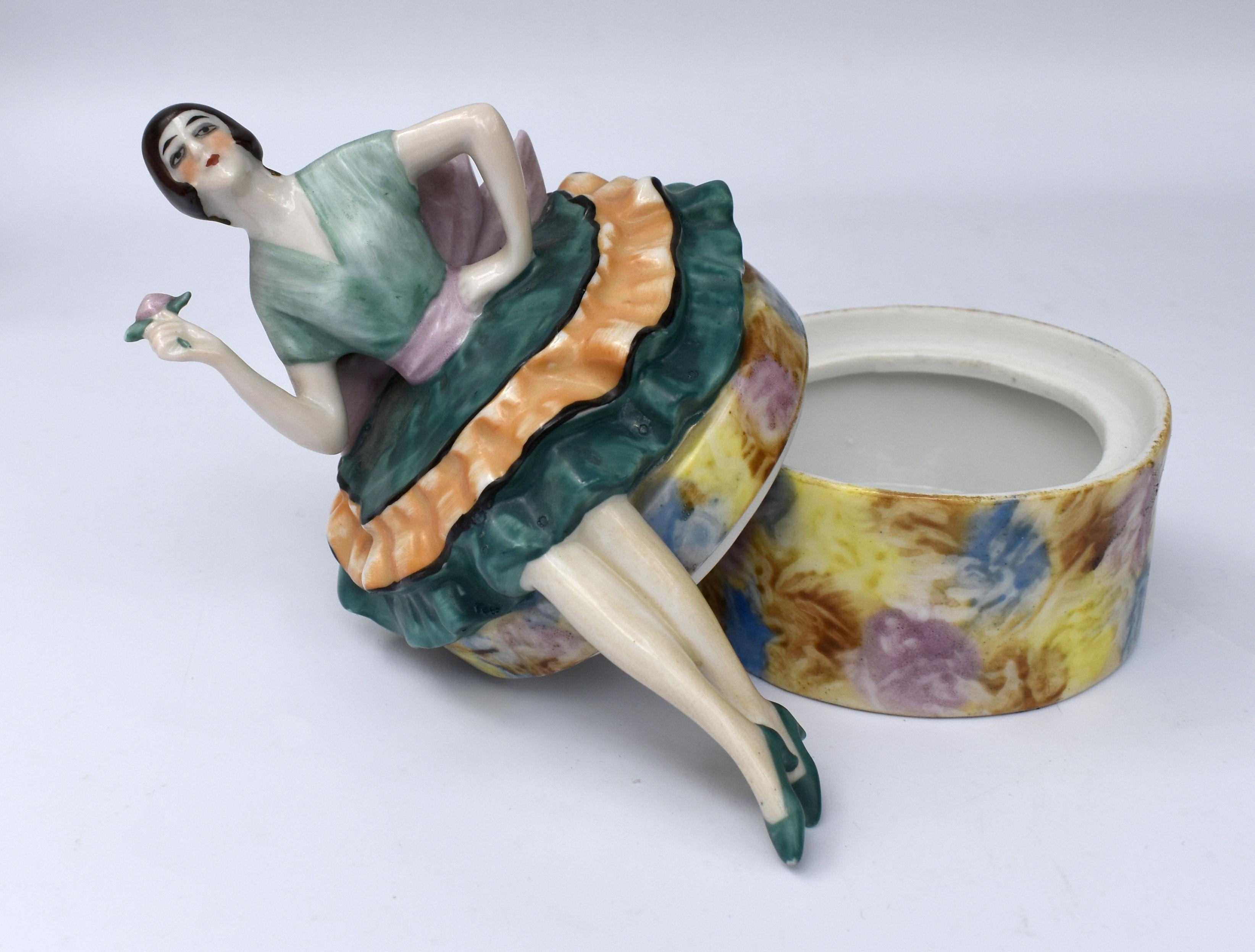 Merveilleuse boîte à poudre en céramique Art déco des années 1930 Cette pièce a la forme d'une boîte à chapeau Art déco avec une jeune fille en vogue assise sur le couvercle et portant une fleur. Magnifique coloration polychrome et détails. Un