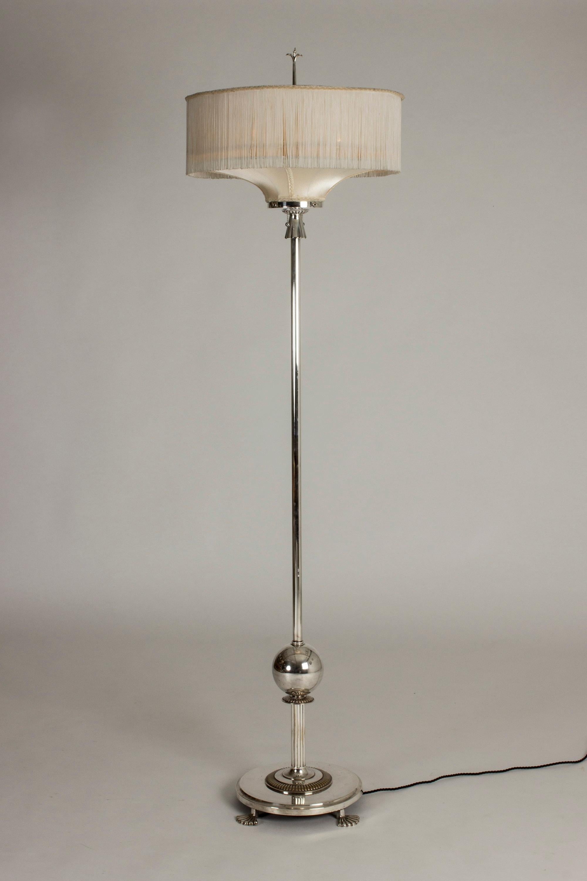 Striking Swedish grace floor lamp by Elis Bergh. Very tall, with a shade dressed with fringe. Beautiful details in the form of a stylized metal lily on top and four lions feet at the base. The two light sources inside can be turned on separately to