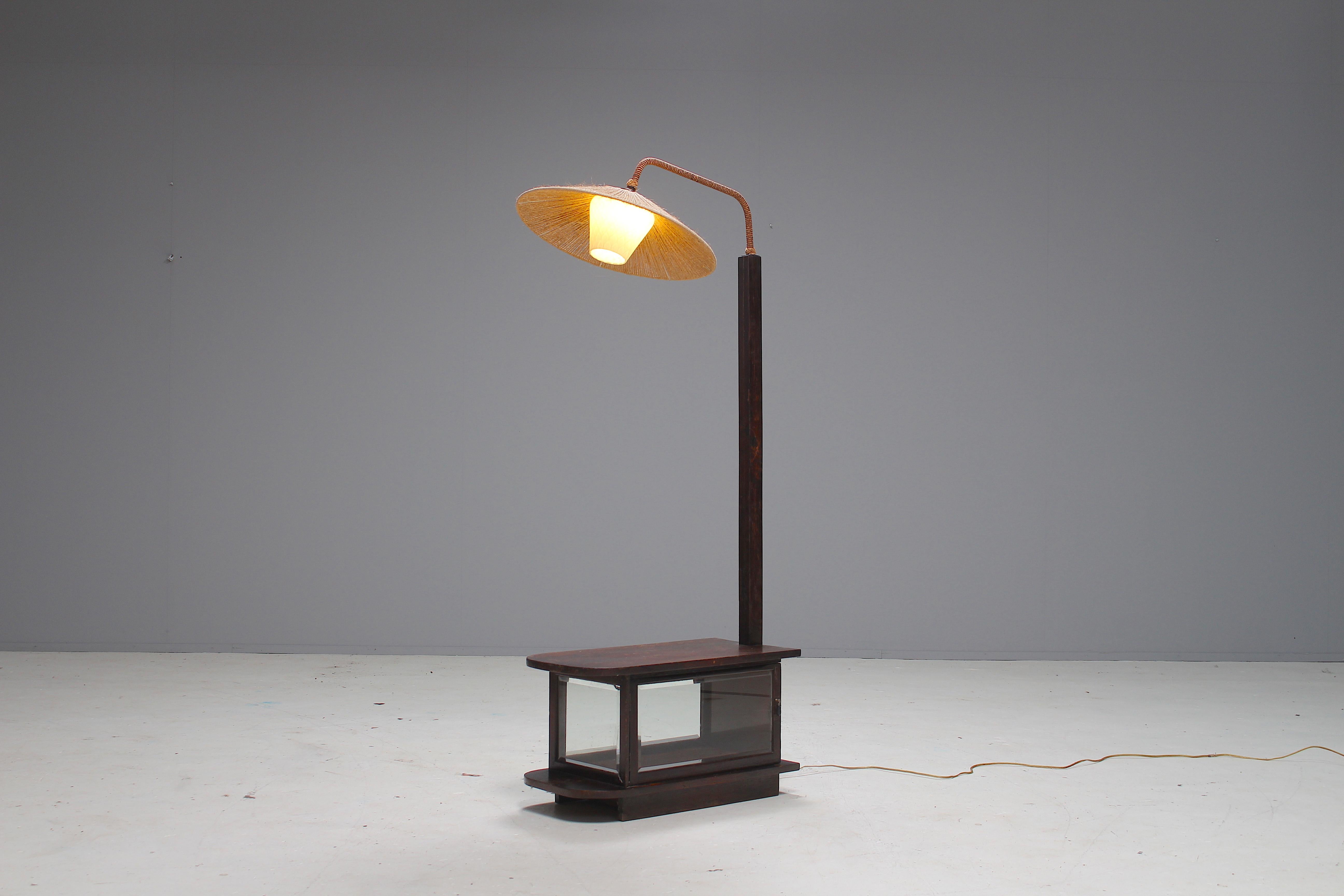 A rare Art Deco floor lamp with a table base and a vitrine.
The lamp shade is made of cord and the diffuser is from opaline white glass.
The vitrine has faceted glass.