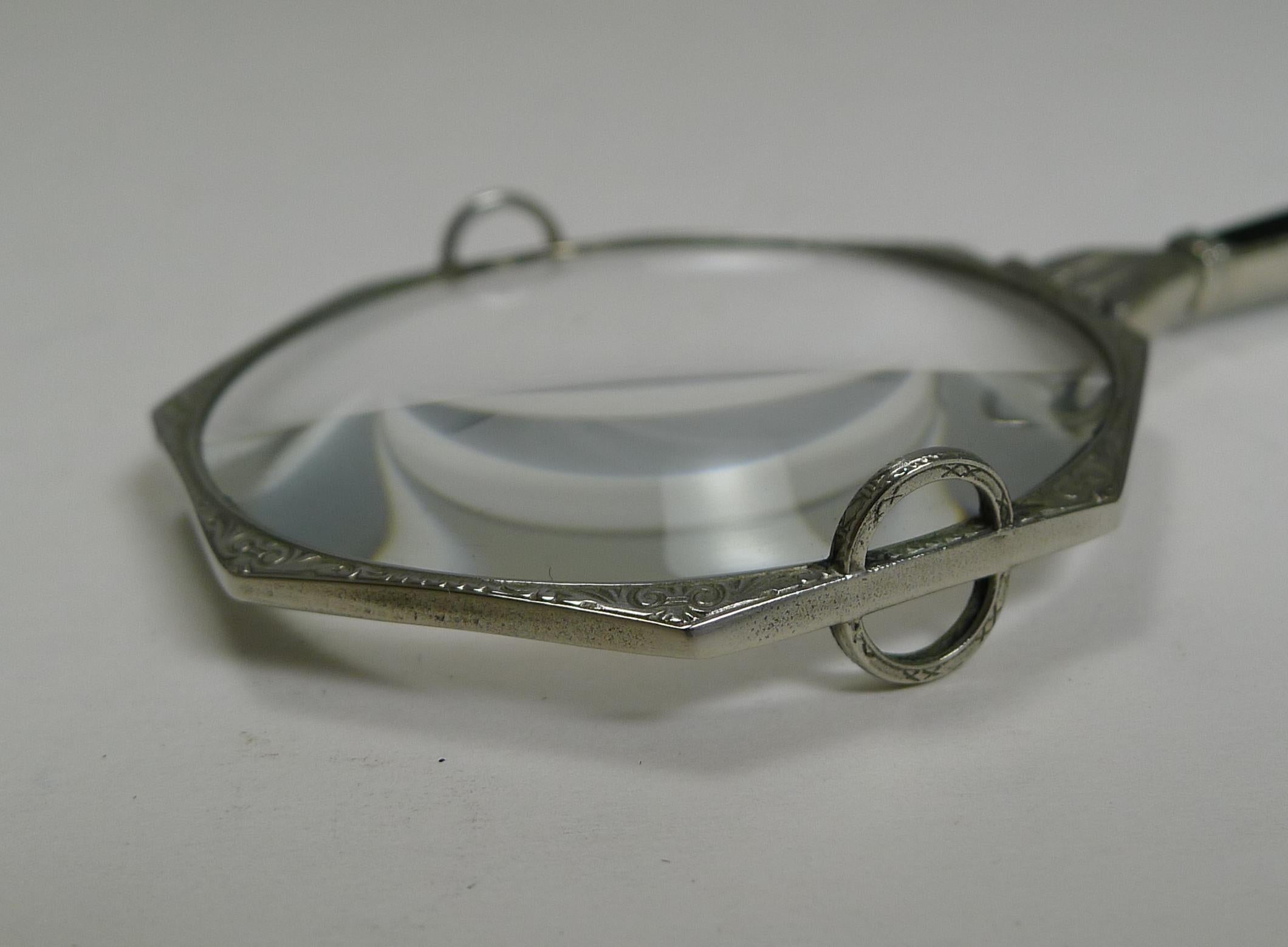 Ein sehr glamouröses Vergrößerungsglas aus weißem Metall (nicht markiert:: aber als Silber getestet) mit eingraviertem Dekor und einem Klappgriff mit einem schwarzen Steineinsatz. 

Auf beiden Seiten des Objektivs befinden sich zwei halbkreisförmige