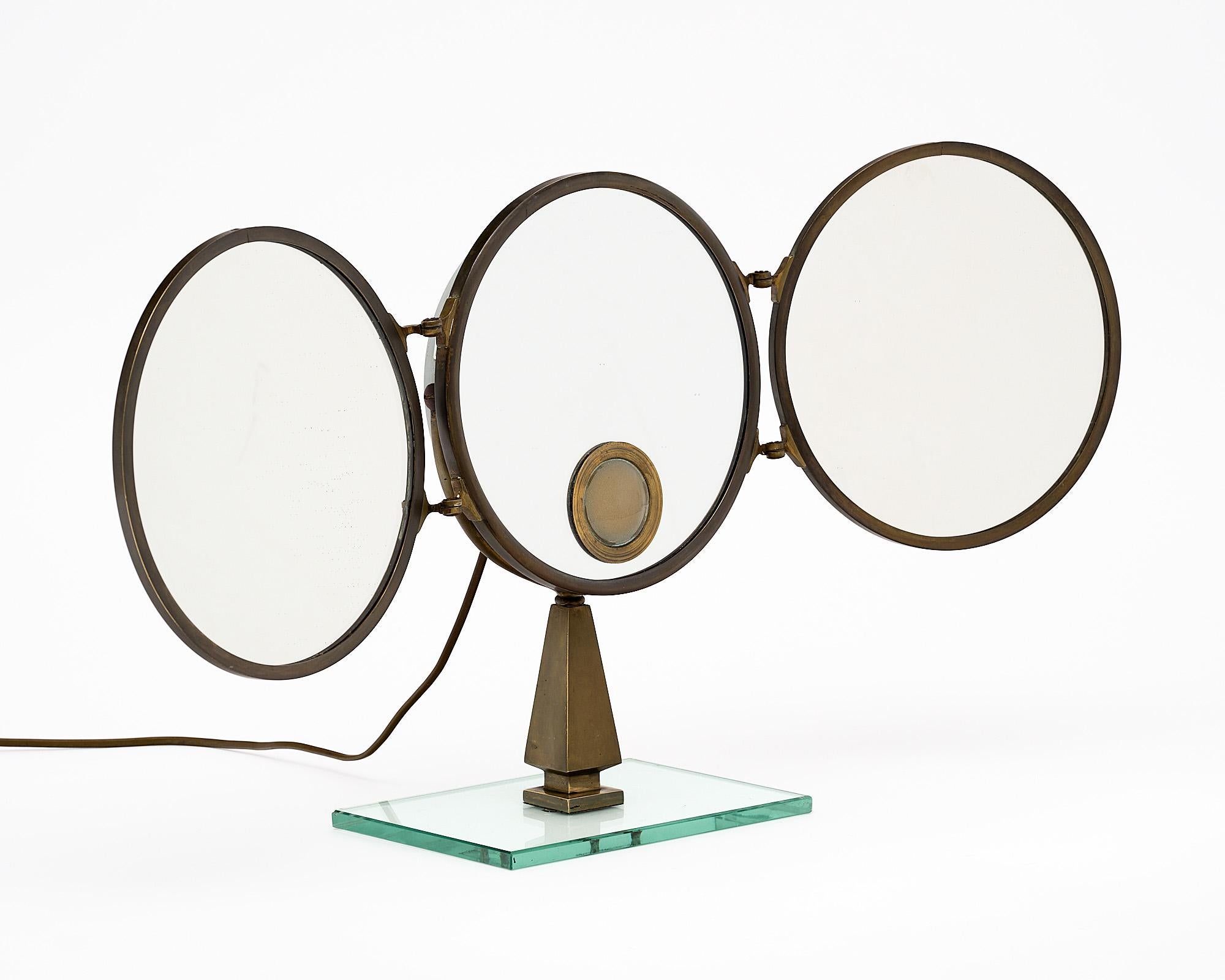 Vanity mirror, French, made of three solid circular brass structures holding mirrors. This triptych piece is articulated. The central mirror features a glass covered cavity equipped with a small socket and bulb. It is supported by a triangular solid