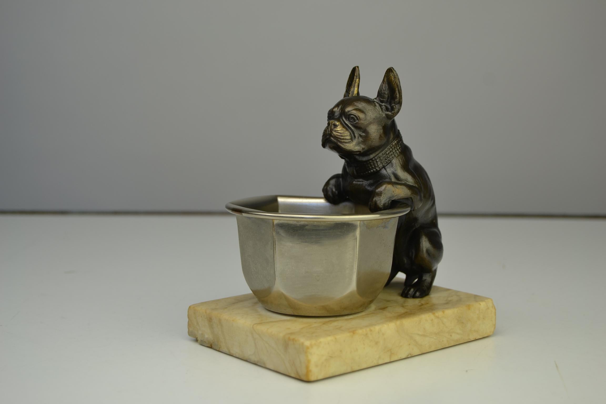 Art Deco French bulldog figurine hanging over a bowl and
mounted on a marble base. 

The bronze colored metal dog sculpture is very detailed.
Look at the snout, the collar and paws.
The metal bowl has a beautiful and rounded hexagonal shape.