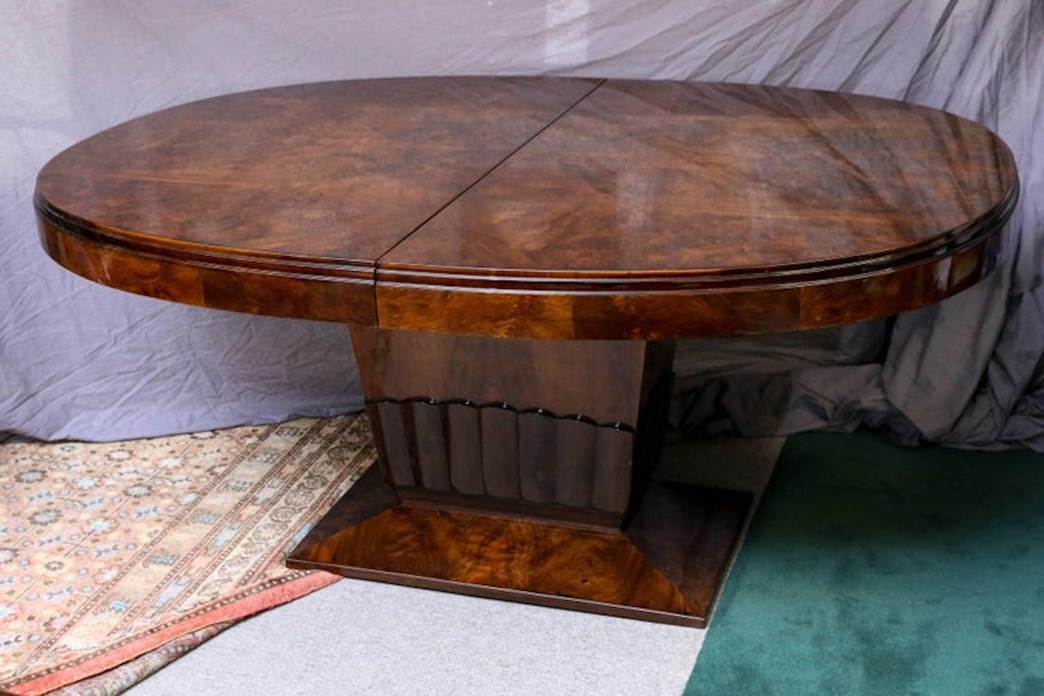 The oval top of the dining room table is displaying the beauty of the burl walnut wood. It is supported by the wide and prominent leg, resting on a rectangular base. Bottom of the supporting leg has decorative “ruffle type” carving. The table has an
