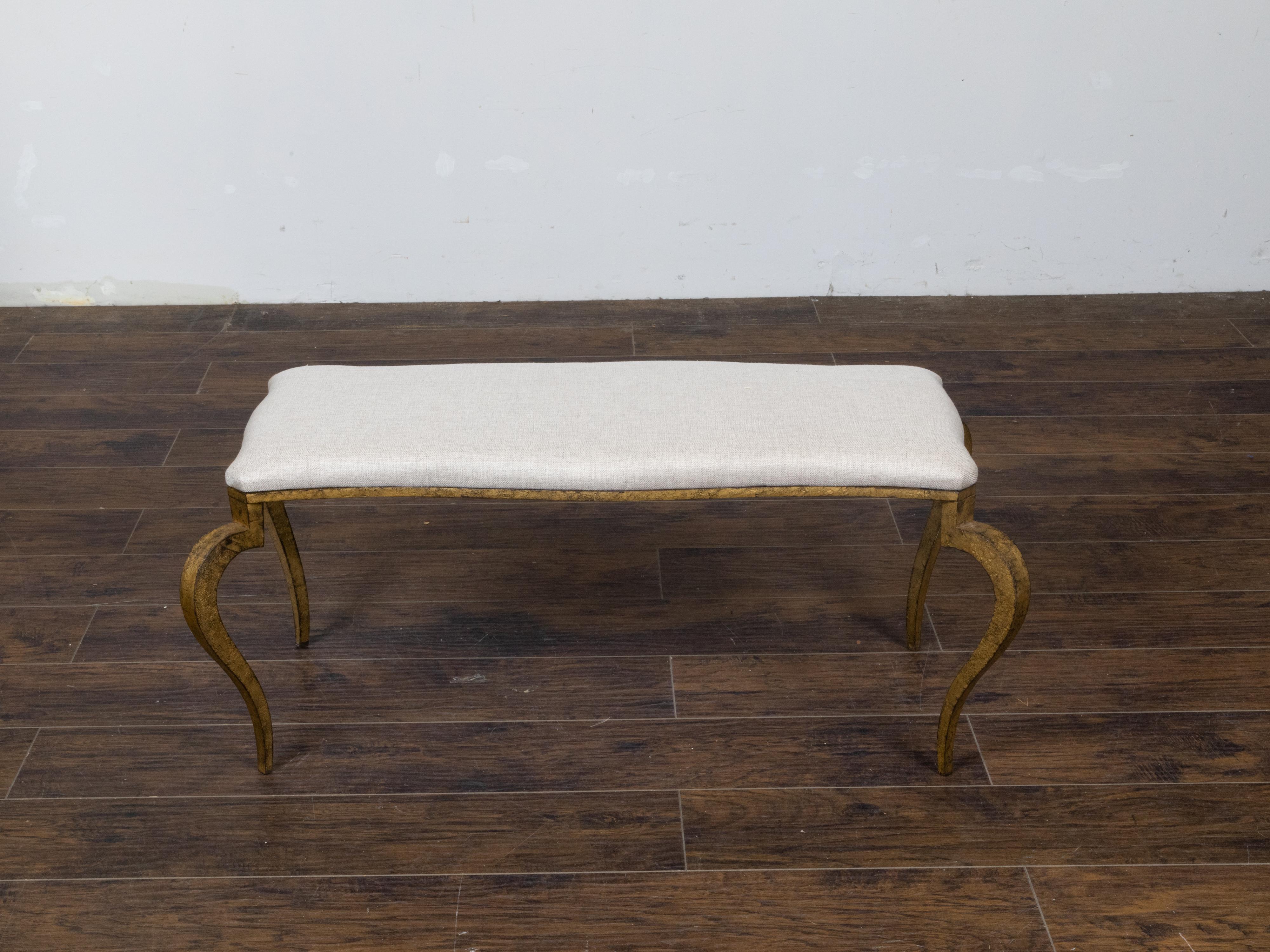 A French Art Deco gilt iron bench circa 1920 with large scrolling and canted legs and rectangular seat newly and professionally reupholstered with a neutral toned linen upholstery. Infuse your interior with a touch of timeless elegance with this