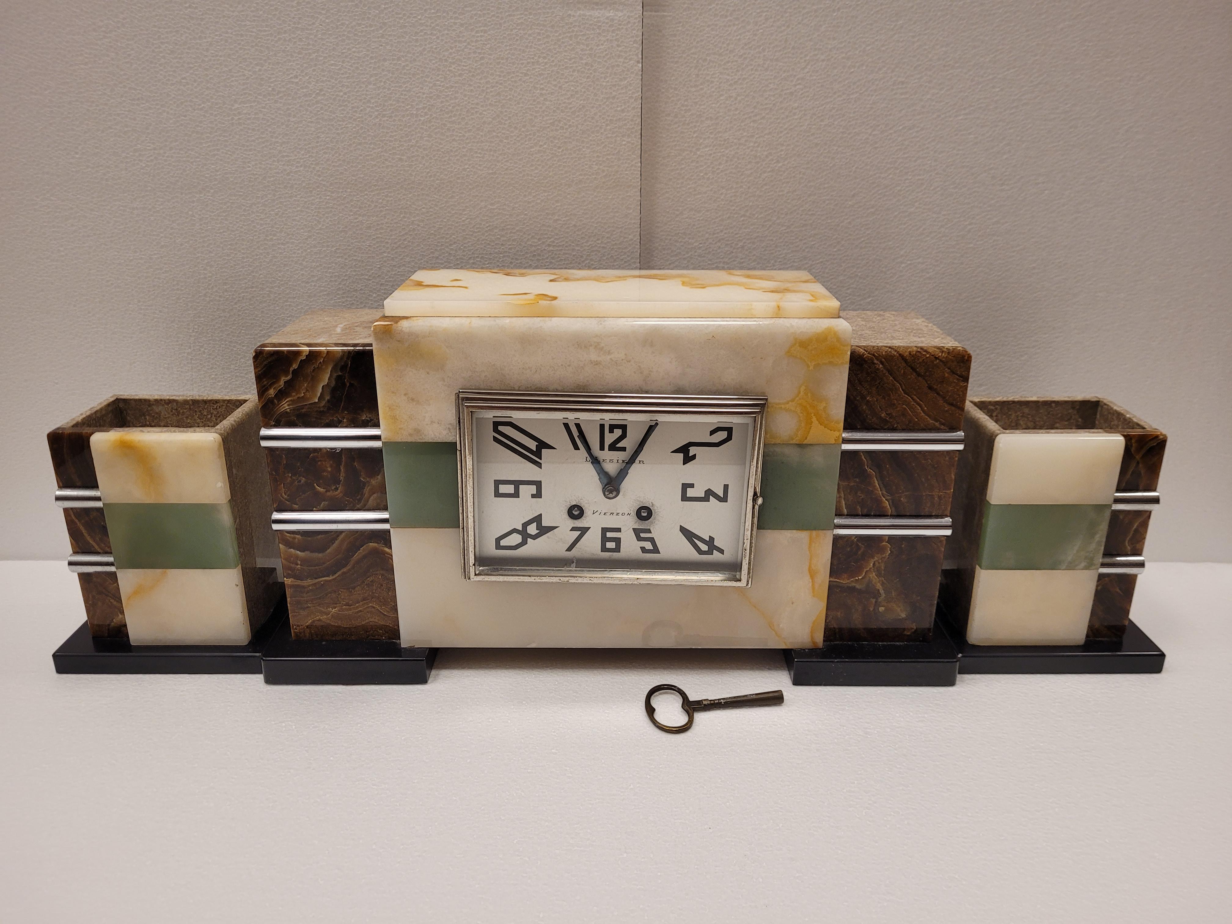 Stunning  French Art Deco style cabinet clock made with stone covering and París machinary. The simplified and symmetrical shape of the body is accompanied by two pieces of marble trim as well. The three pieces are enriched on the front with steel