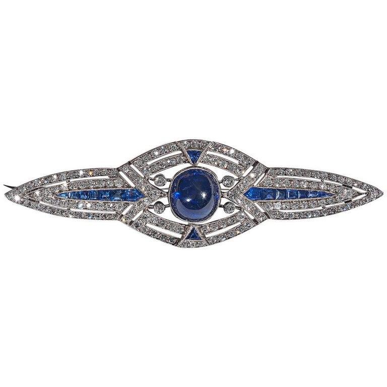 Of geometric outline, the oval cabochon principal sapphire stone bordered by a diamond clusters, radiating two lines of calibre cut sapphires to the sides and single triangular sapphires at the top and bottom, millegrain set.
Mounted in