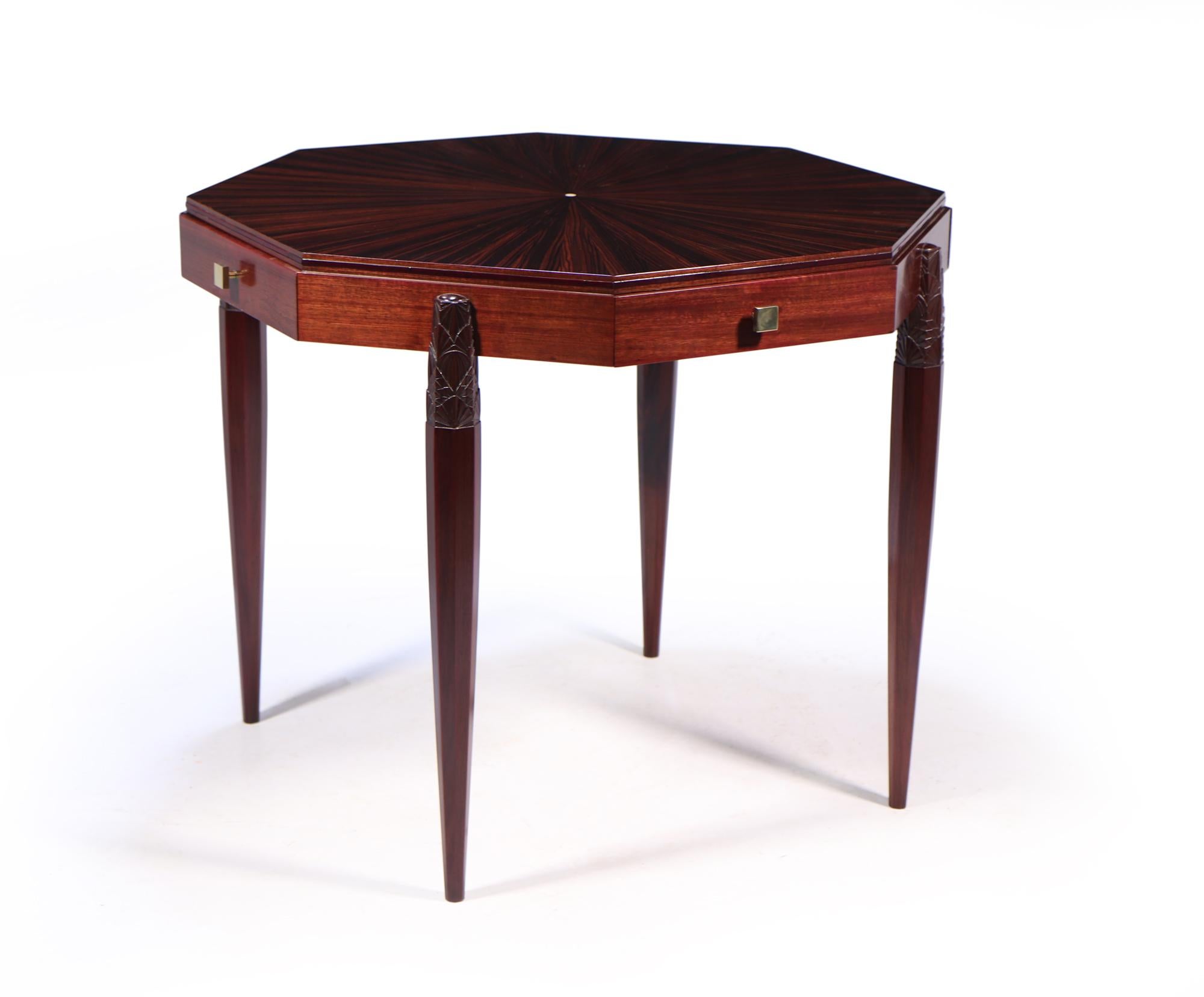 An octagonal shaped games table with four drawers with brass square handles and octagonal shaped legs with crisply carved art deco pattern to the top of the leg. The table is a solid mahogany frame with a segmented macassar ebony top and a mother of