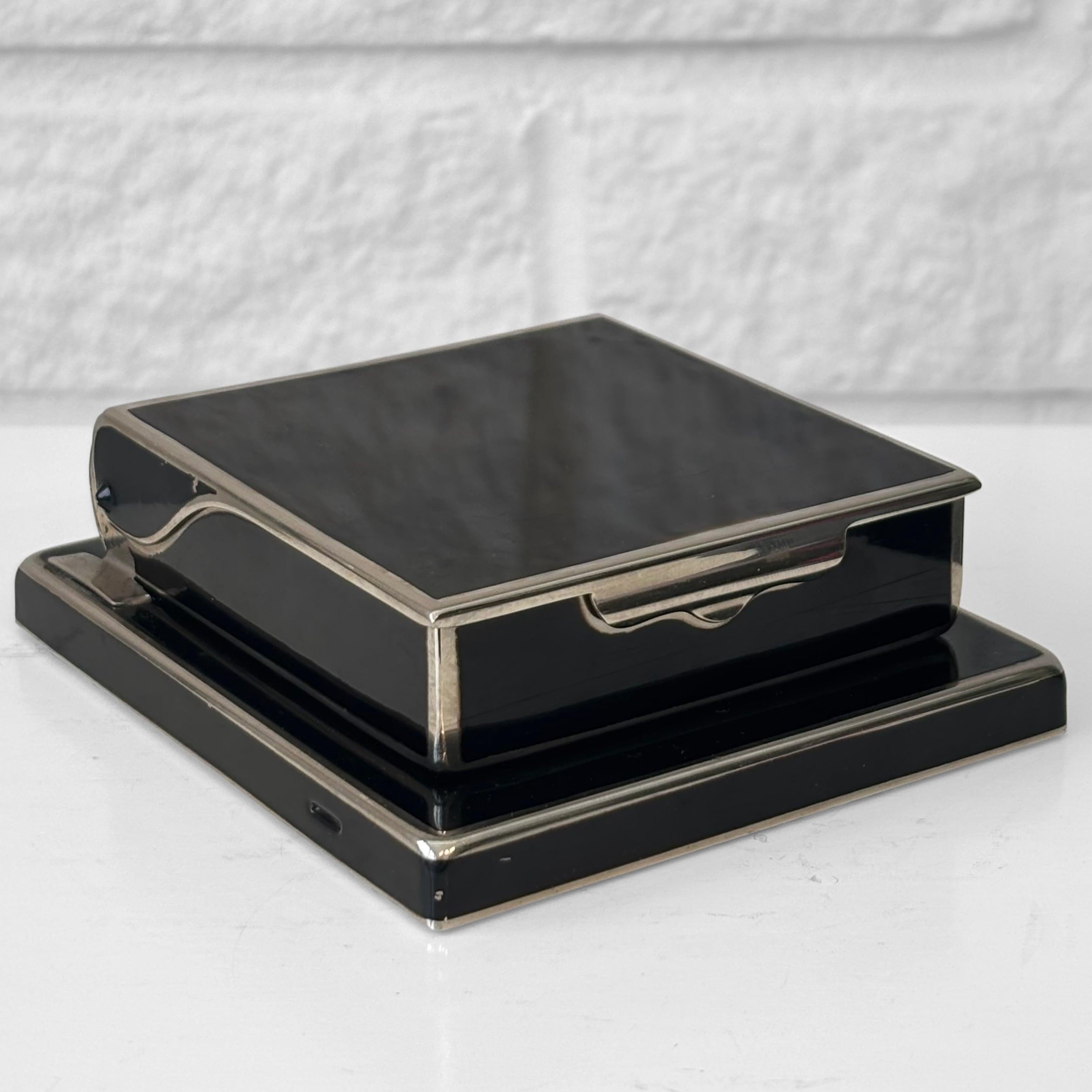 A small cigarette dispenser crafted from enamel steel, likely produced by the German manufacturer Rowenta in the 1930s. It consists of a square base and a box-shaped dispenser that reveals a cigarette when placed in an upright position. Engraved