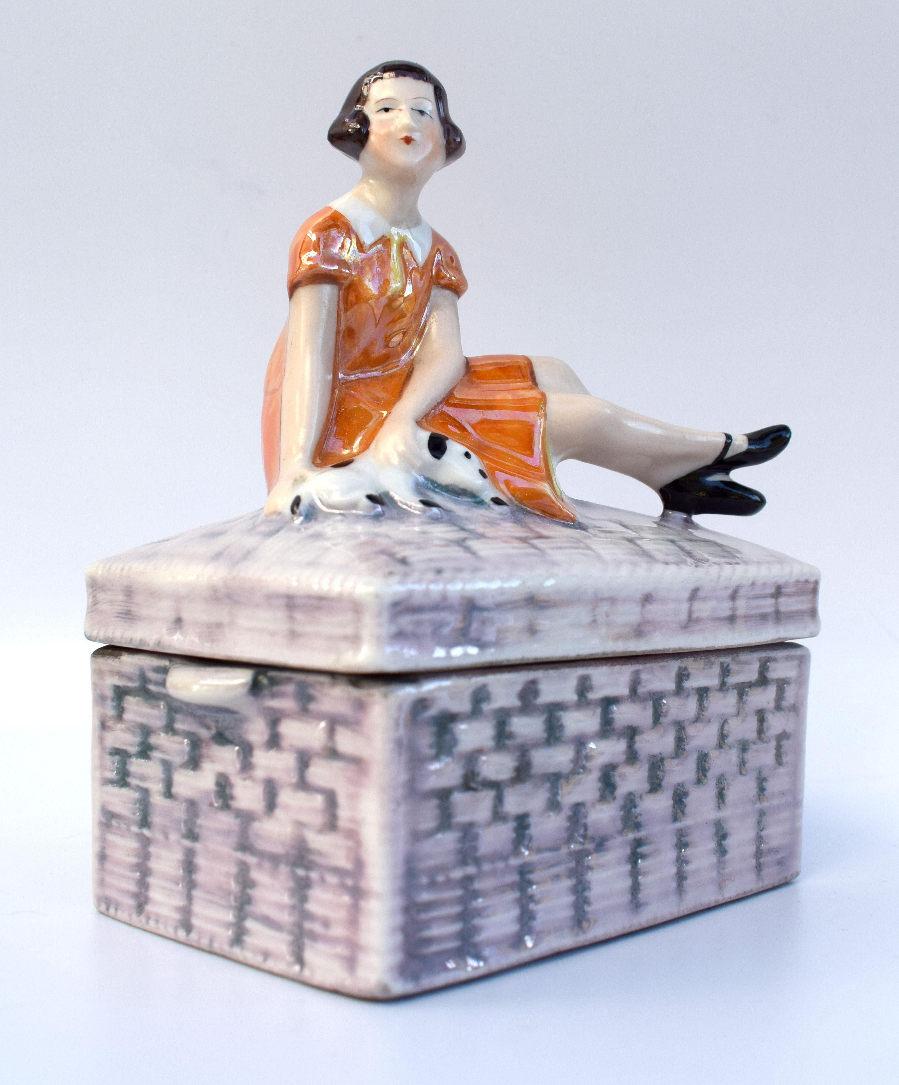 Striking and quite rare is this 1930s Art Deco porcelain powder box with lustre finish which depicts a young girl sat on a wicker trunk stroking her puppy dog which lays next to her. Beautiful coloring and detailing. A rare design and highly sought