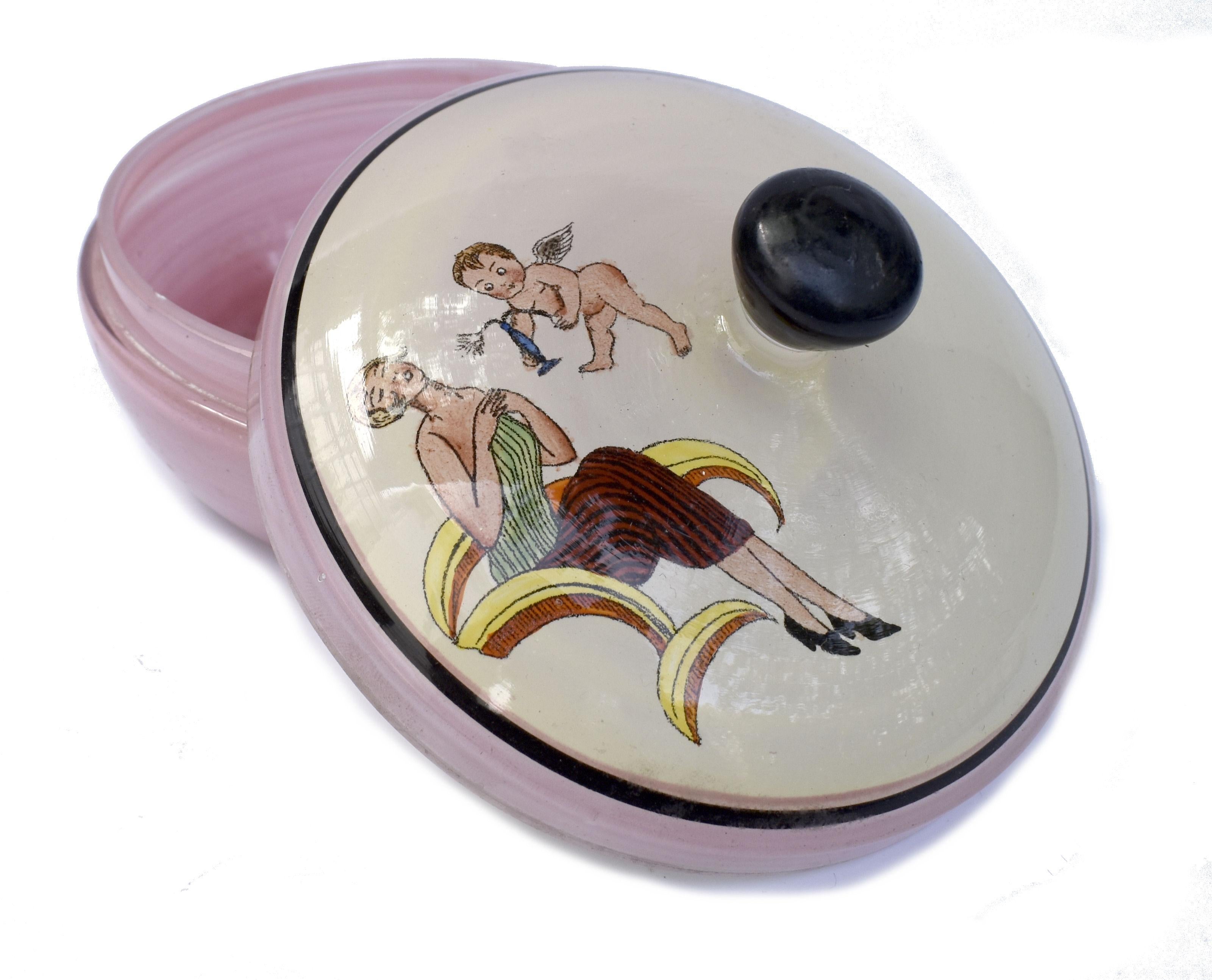 Nous vous proposons ce charmant bol à poudre en verre rose Art Déco des années 1930. Sur le couvercle, une jeune fille à la mode de l'époque est représentée à côté d'un cupidon / chérubin volant tenant un arc et une flèche. Très bel article et