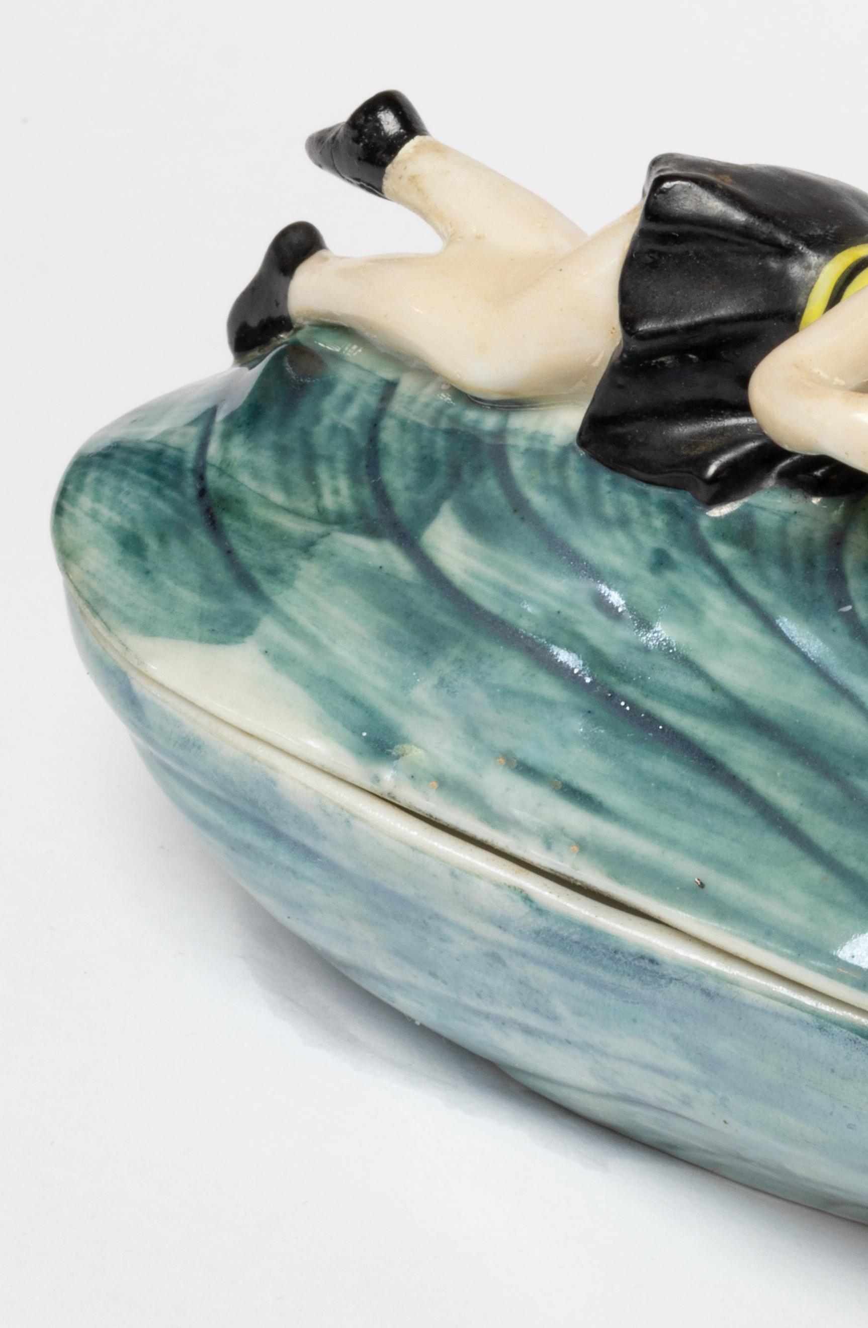 An Art Deco porcelain powder box featuring a reclining bathing beauty atop a mussel shell, crafted by Goebel in Bavaria, 1920.

This rare and collectible piece perfectly embodies the elegance and playful charm of the 1920s. 

Molded in glazed