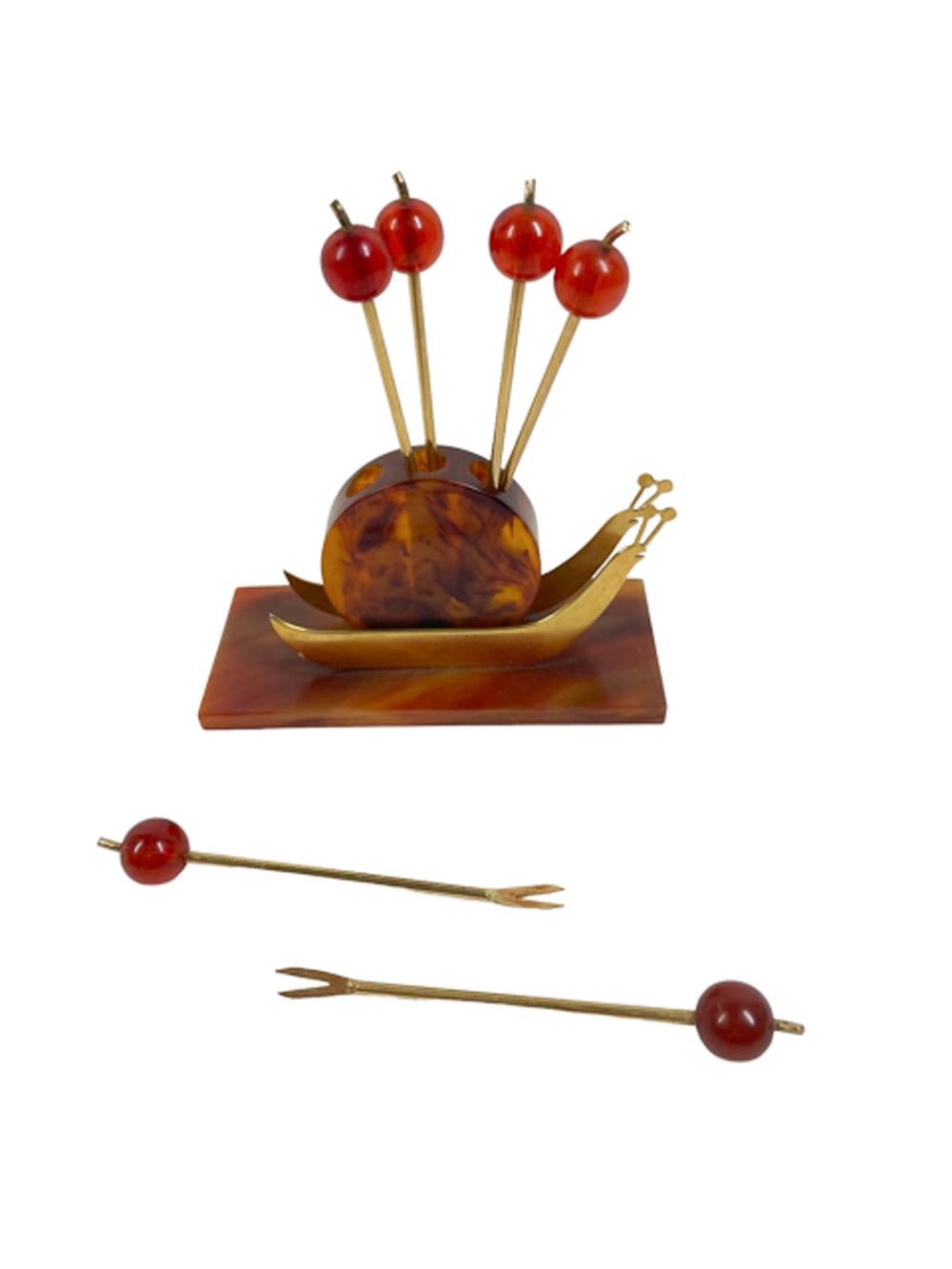 Art Deco cocktail pick set with six gold toned picks with forked tips and translucent red berry tops in a snail-form stand of faux tortoiseshell Bakelite and gild toned metal.