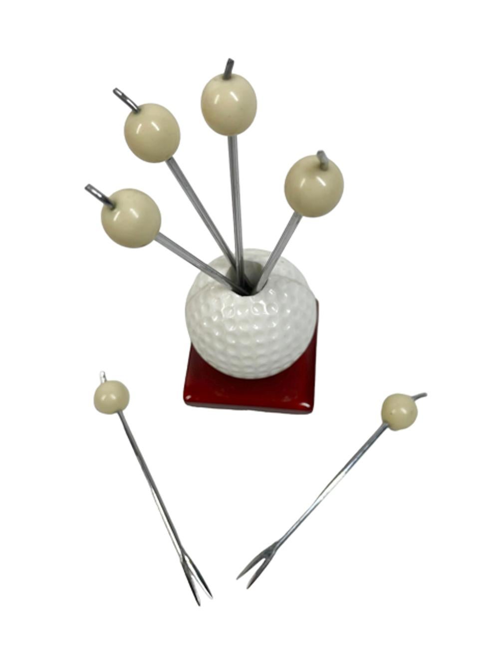 Set of six white ball topped forked chrome cocktail picks in a white plastic golf ball holder with a square translucent apple juice colored Bakelite base. 