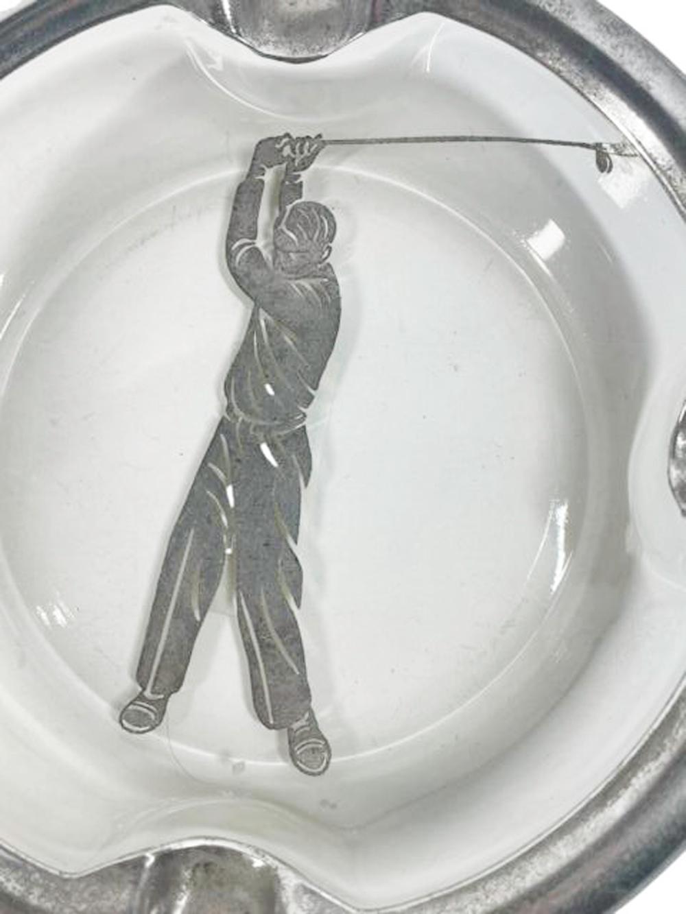 Cenicero de cristal plateado de forma circular, con el borde plateado en relieve y cuatro apoyos para puros/cigarrillos centrados en un golfista plateado en pleno swing sobre fondo transparente.