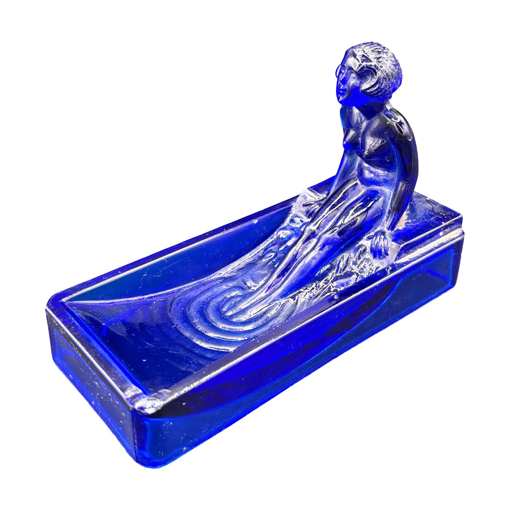 Cette réédition des années 1980 d'un porte-savon/plateau à anneaux en verre bleu Heinrich Hoffman de 1927 Art déco représente une femme nue se baignant dans une baignoire.

Cette réédition est signée et fabriquée par Sarsaparilla à New York. Elle