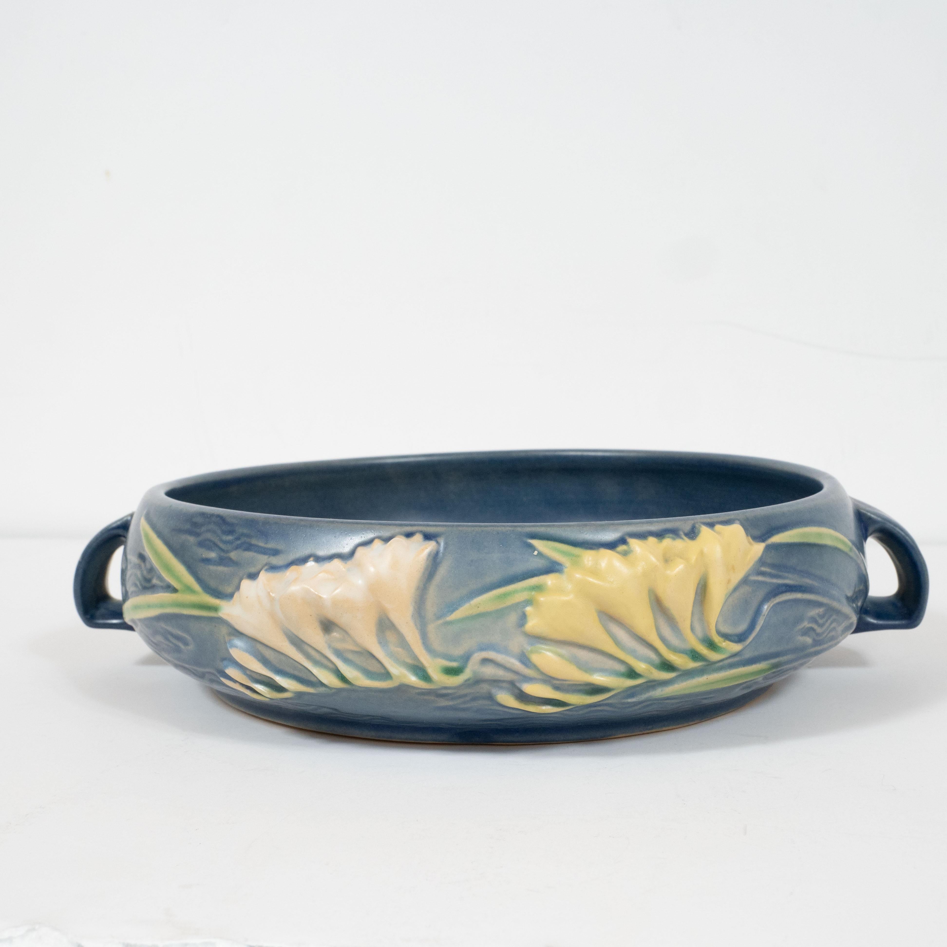 This elegant Art Deco bowl was realized by the esteemed maker Roseville Pottery in the United States, circa 1930. It features a bowed circular body in a rich indigo hue with lily of the valley flowers presented in relief and sculptural handles.