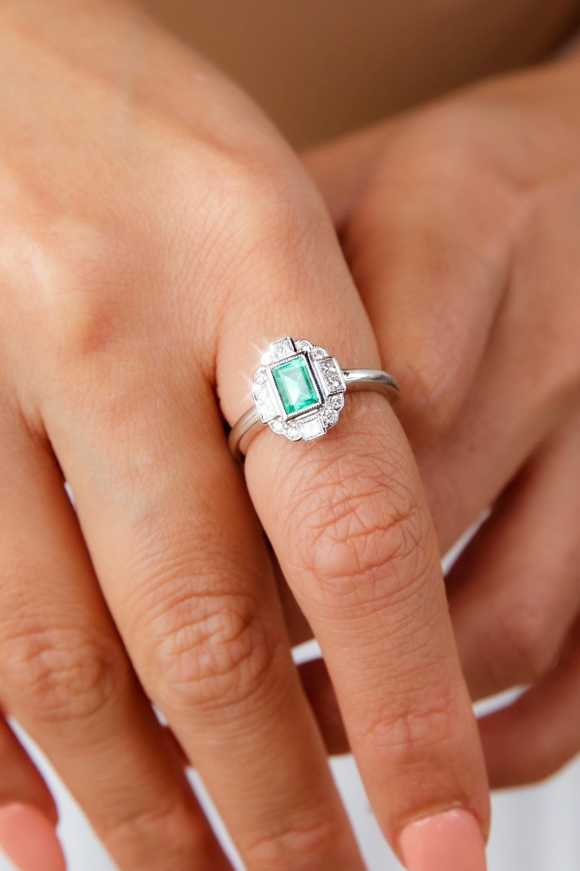 Lovingly forged in 18 carat white gold, this enchanting art deco inspired contemporary ring features a lovely bright green emerald at the centre of a captivating millegrain border of shimmering princess cut and round brilliant cut diamonds. This