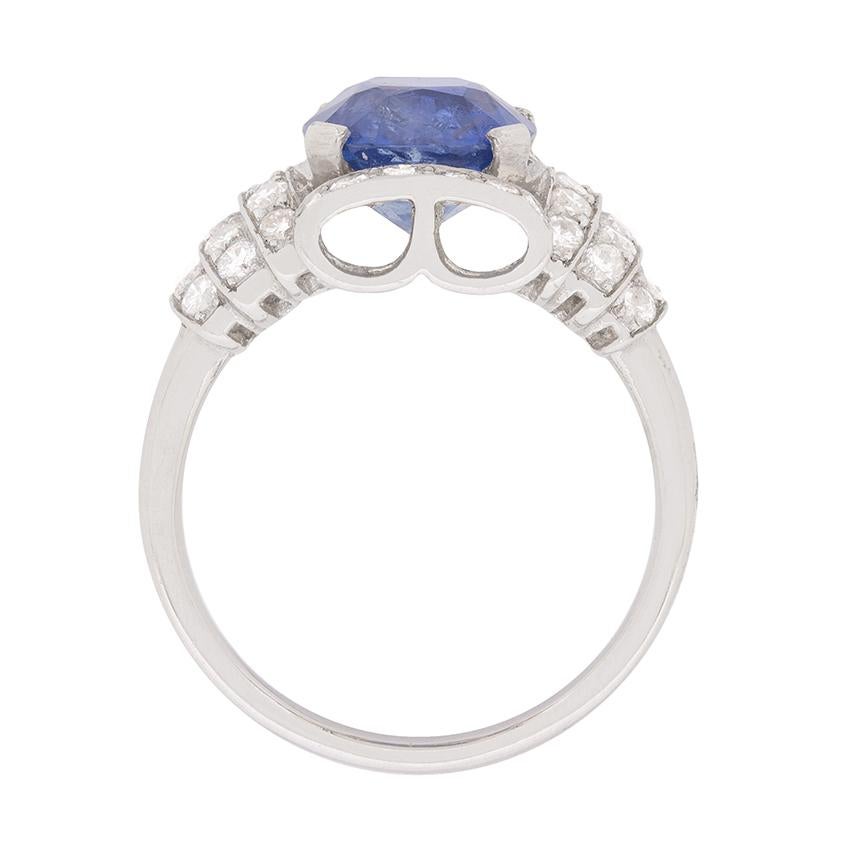 A strong Art Deco Style influence is evident in this fabulous circa 1950s sapphire and diamond ring. This wonderful mid-century piece showcases a stunning 2.76 carat, transparent blue, cushion cut Burmese sapphire amid a shimmering grain set border