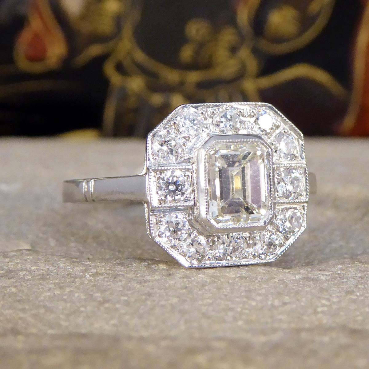 This beautiful contemporary ring has been hand crafted from Platinum in an Art Deco style. It features a centre Emerald Cut Diamond in a millegrain edge setting weighing 1.00ct with a slight colour draw giving it an almost champagne light lemon