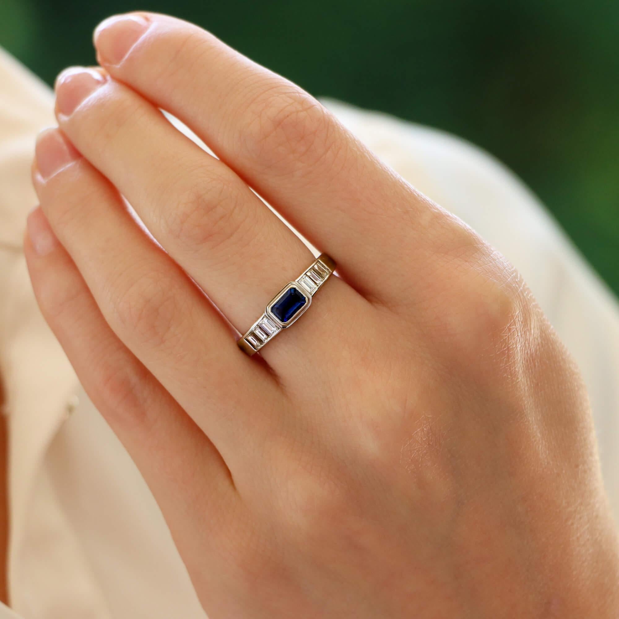 A highly unique Art Deco inspired sapphire and diamond ring set in 18k white gold.

The ring is centrally set with a 0.70 carat emerald cut blue sapphire which is securely rub over set in a platinum. To each shoulder are three graduating emerald cut