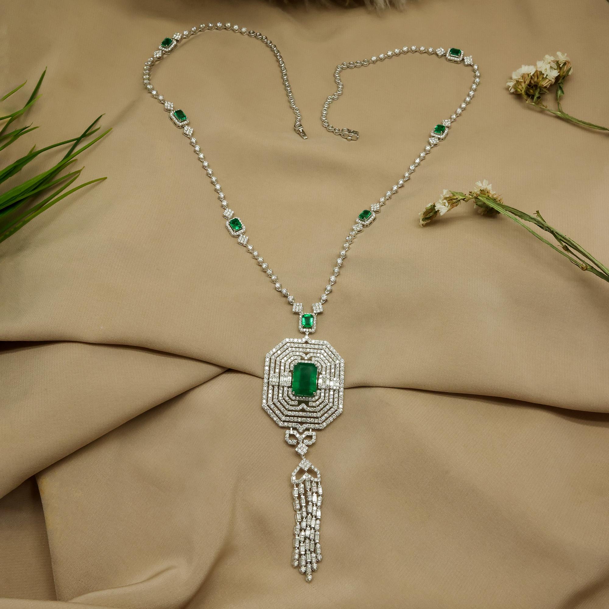 Questa collana a goccia in oro bianco 18 carati con smeraldo dello Zambia, ispirata al periodo Art Deco, è caratterizzata da uno smeraldo con cinque strati di diamanti a raggiera che formano un ciondolo con linee di diamanti che penzolano da una